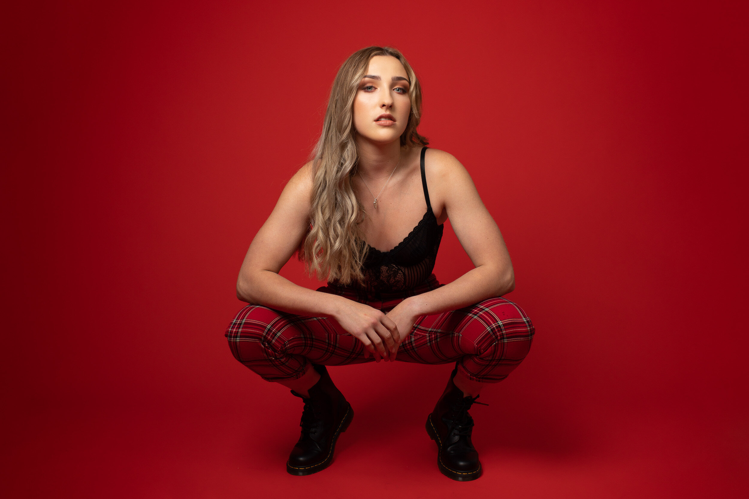 Person squatting in front of red background wearing red plaid pants and black top.