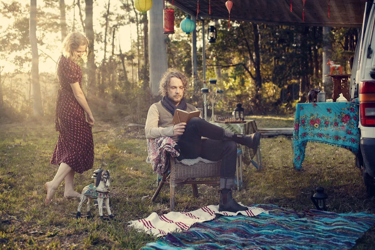 Man reading outdoors next to a woman and a decorated table with hanging lanterns, blankets, and forest backdrop.