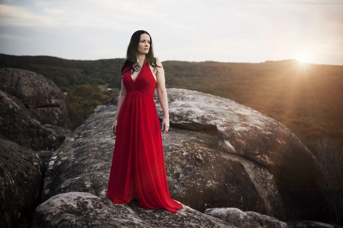 Woman in a red dress standing on rocky terrain with a scenic landscape and sunset in the background.