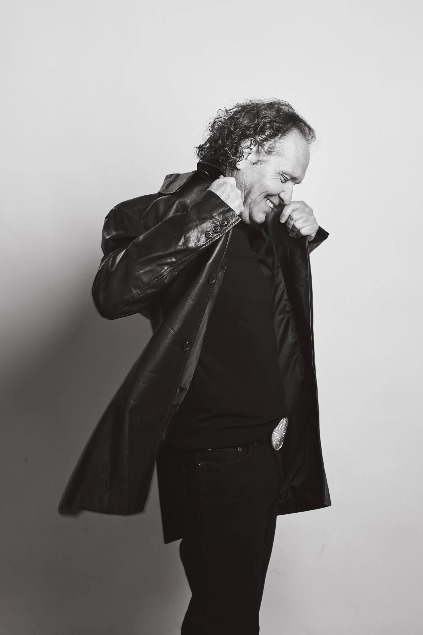 Black and white photo of a person in a leather jacket and black outfit, smiling and adjusting the collar.