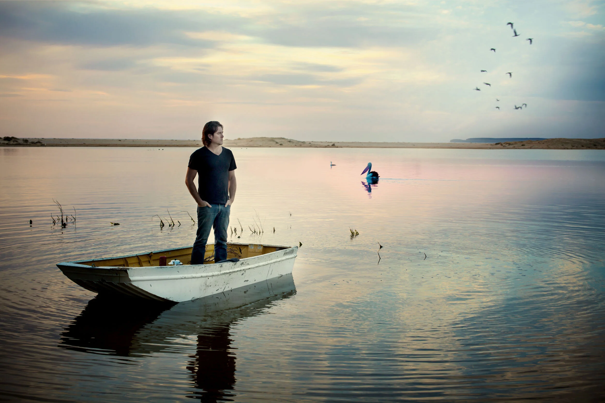 Man standing on a small boat in a calm lake at sunset, with birds and reflections on the water.