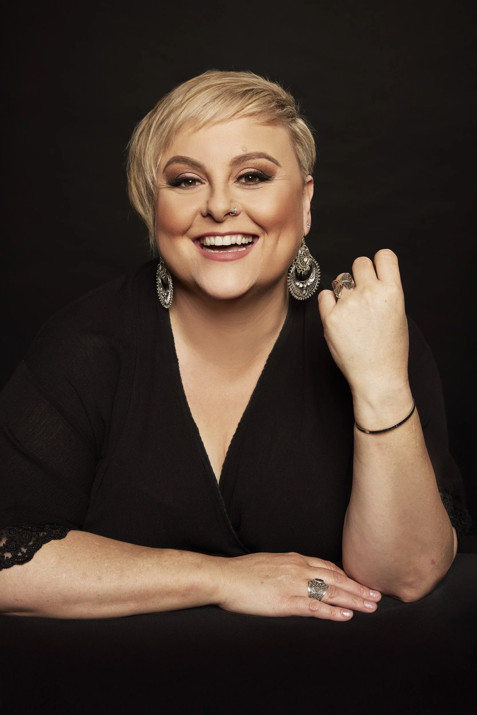 Lyn Bowtell smiling with short blonde hair, wearing large earrings and a nose ring, in a black outfit against a dark background.