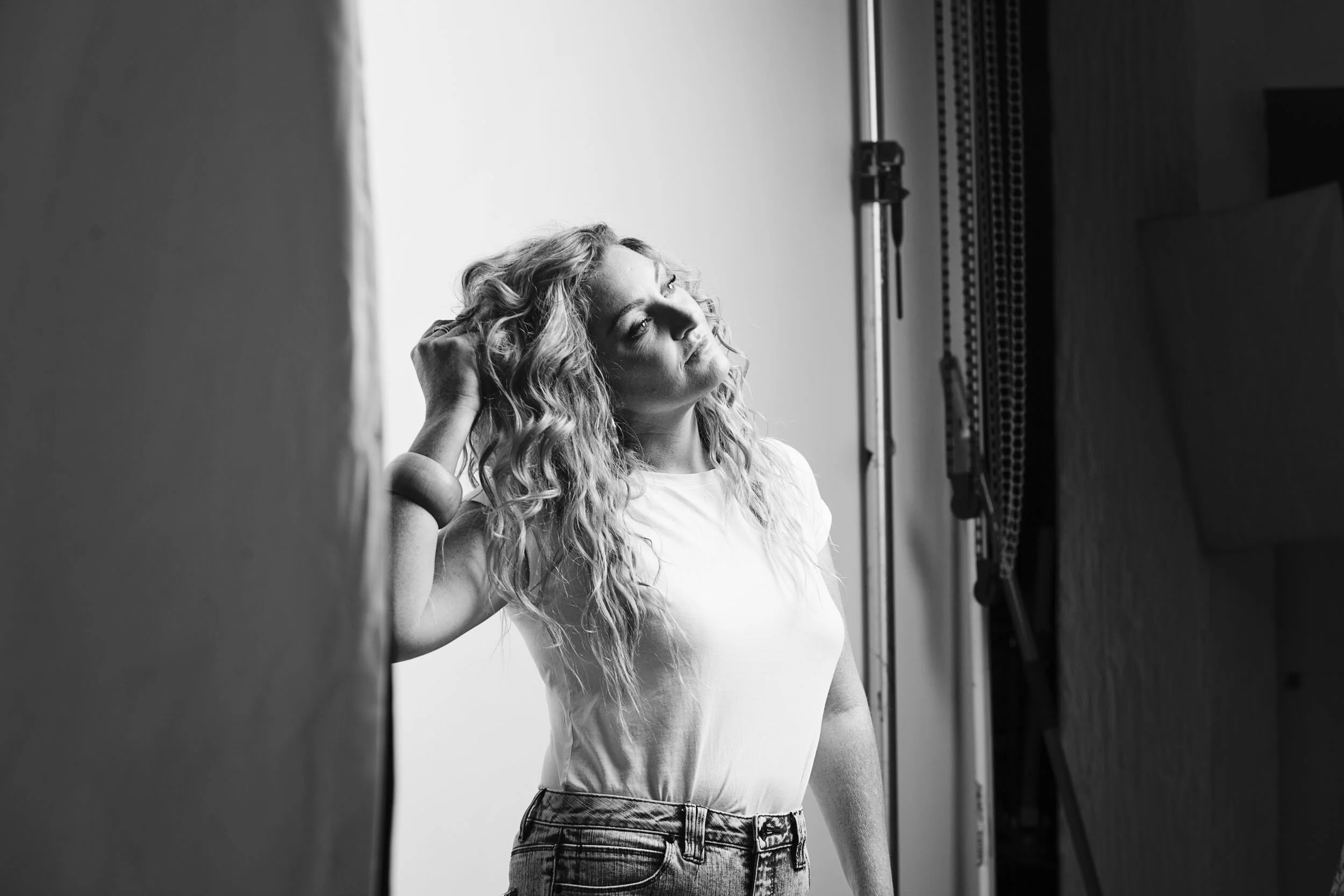 Black and white photo of Nat Henry with curly hair touching her head, wearing a white shirt and jeans, standing in a studio setting.