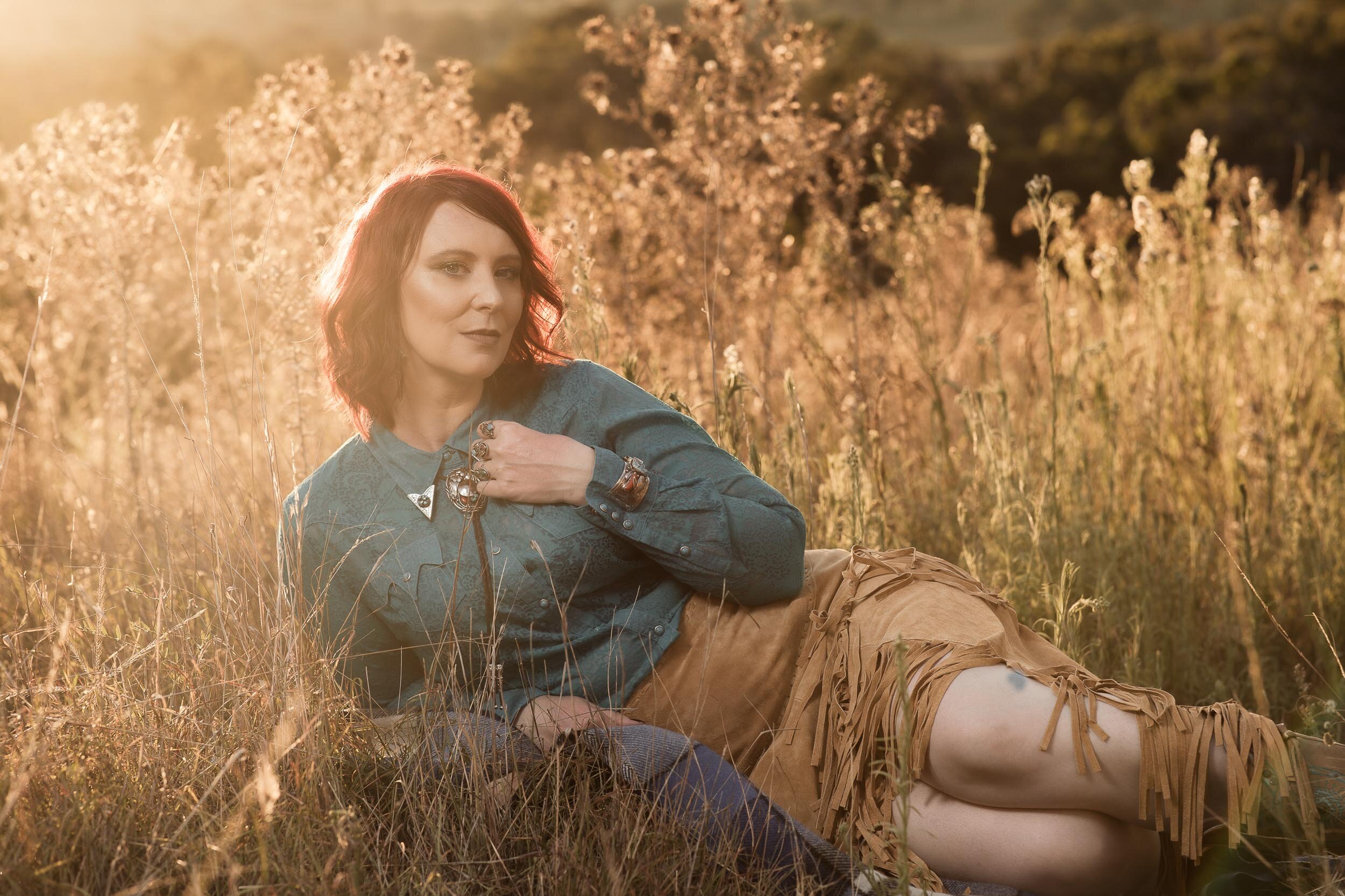 Person lying in field, wearing blue shirt and fringed tan skirt, with sunlight in background.