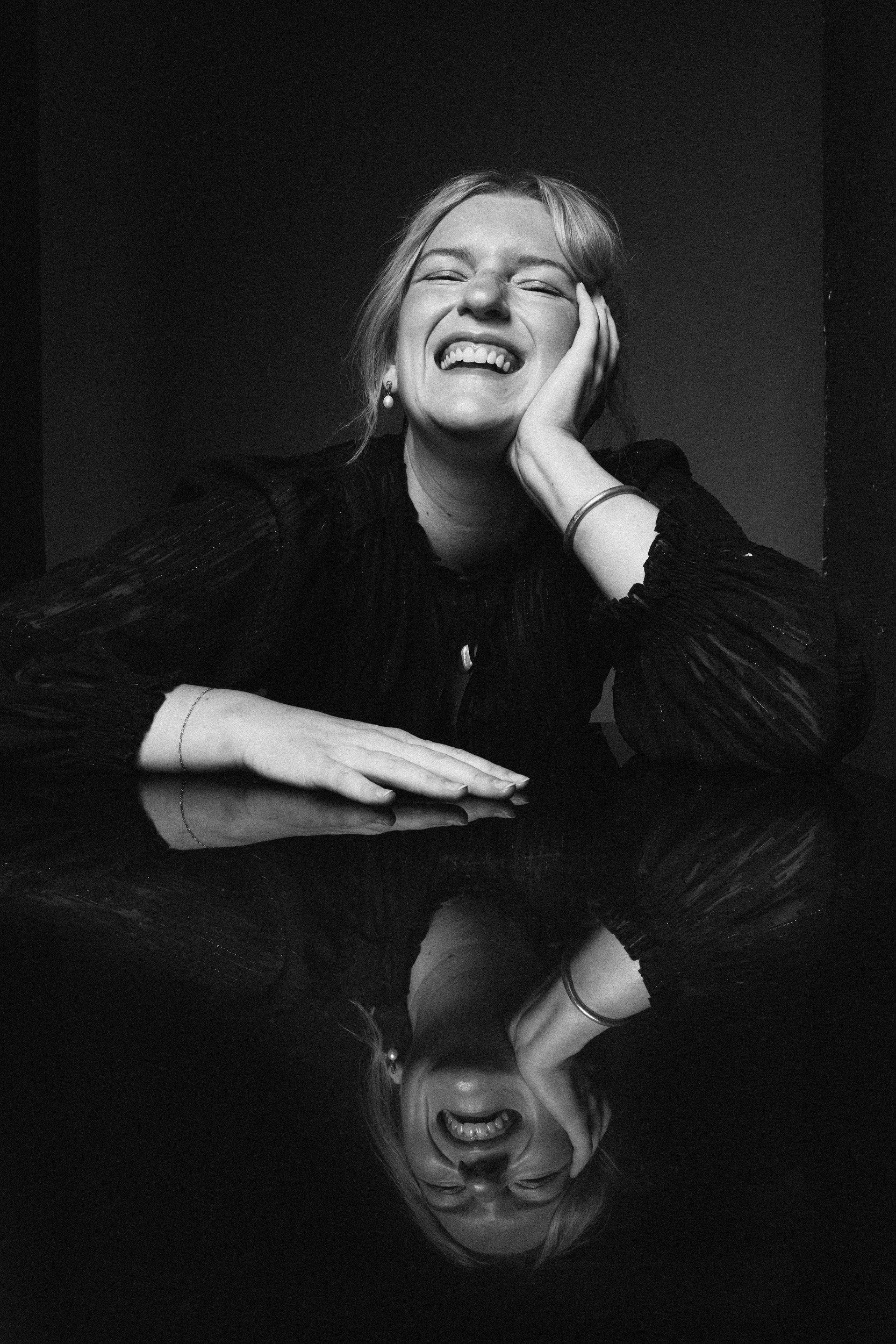 Black and white photo of a smiling woman resting her head on her hand, with her reflection visible on a glossy surface.