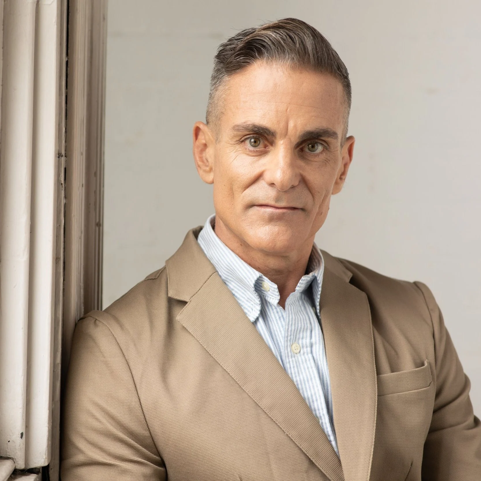 Man in brown suit leaning against a wall, wearing a light blue striped shirt.
