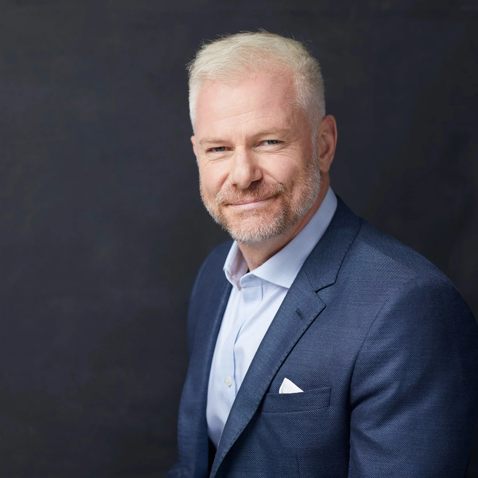 Man with short white hair and beard in a blue suit, smiling against a dark background.