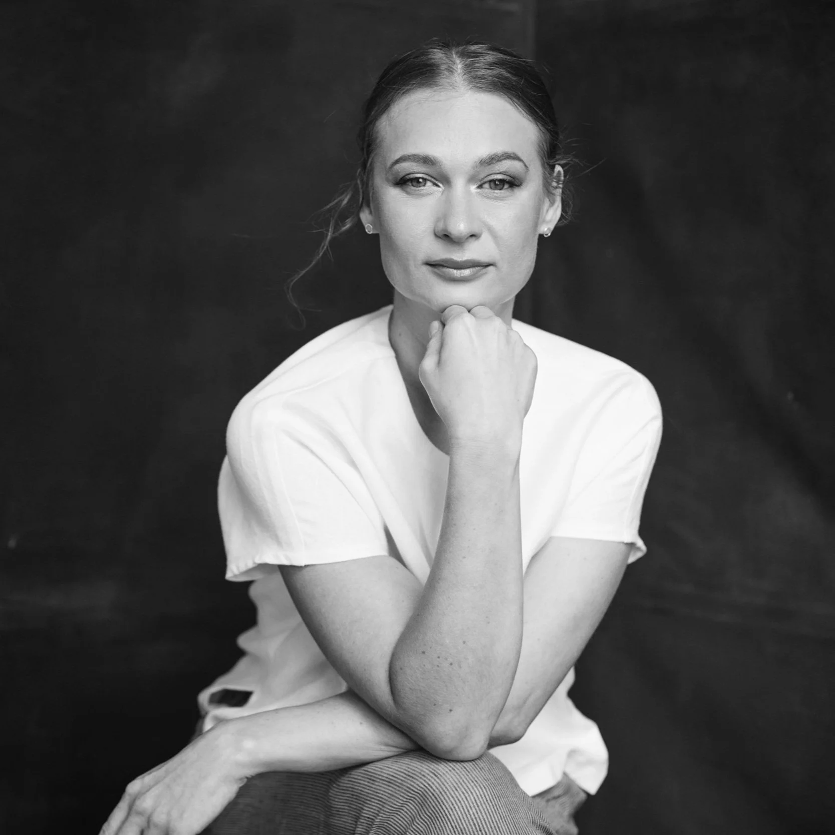 Black and white portrait of a woman in a white shirt, resting her chin on her hand, looking at the camera.