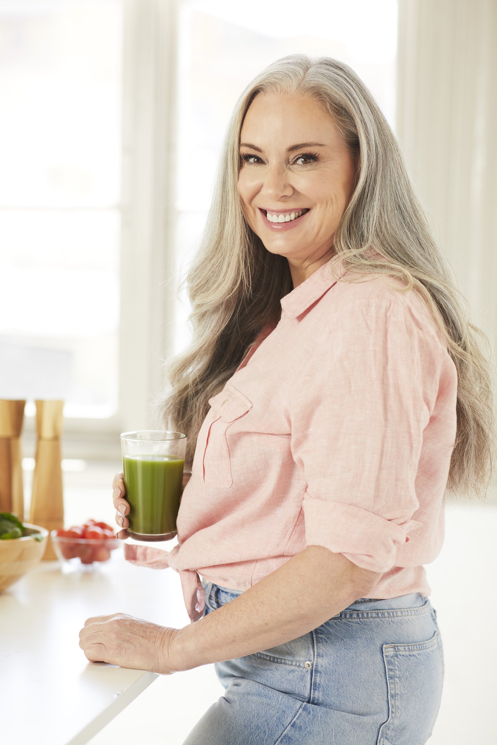 Smiling older woman with long gray hair holding a glass of green juice in a bright kitchen.