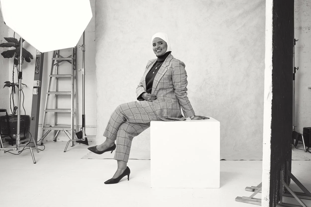 A black and white photo of a person sitting on a studio prop, wearing a checkered suit and hijab. The setup includes a studio light, ladder, and light background. The person is smiling and posing confidently.
