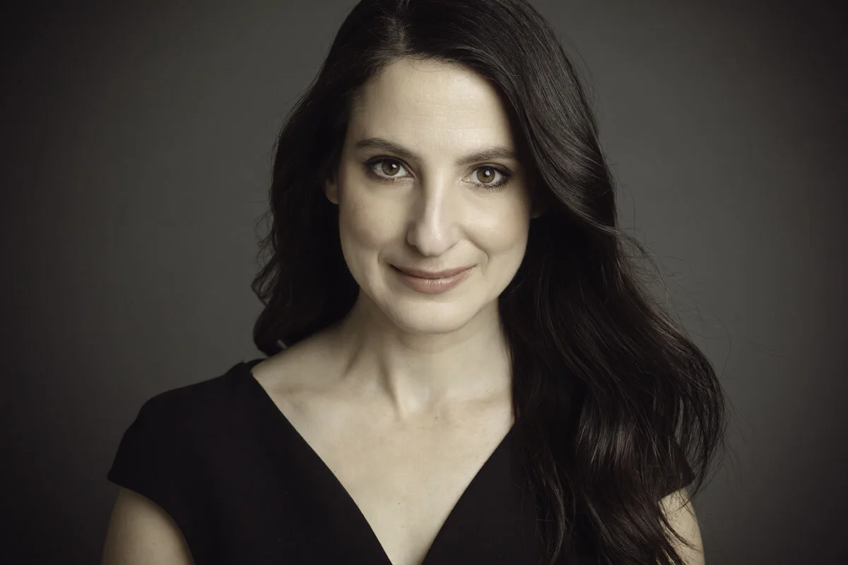 Portrait of a woman with long dark hair smiling slightly, wearing a black top, against a dark background.