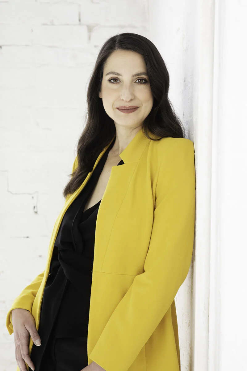 Woman in yellow blazer leaning against a white brick wall, smiling.