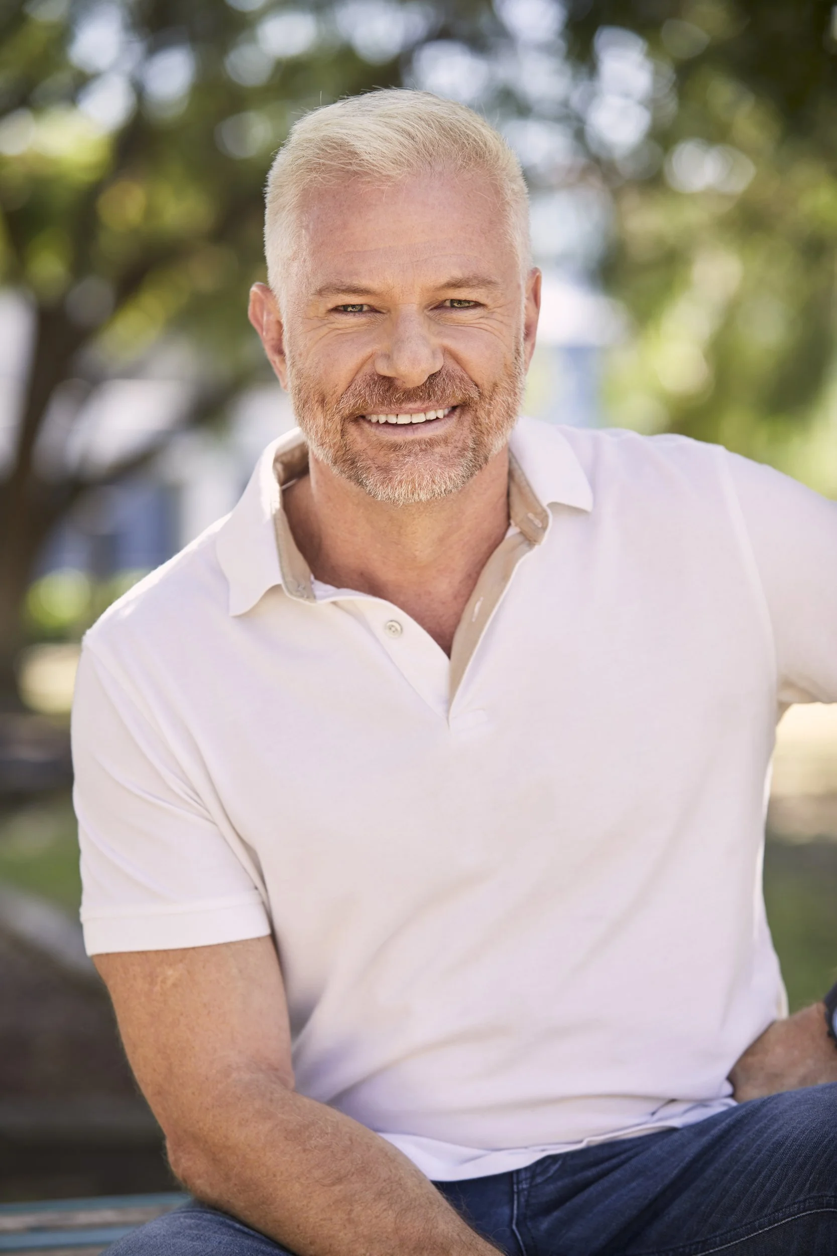 Smiling man with short blond hair and beard wearing a white polo shirt, seated outdoors.