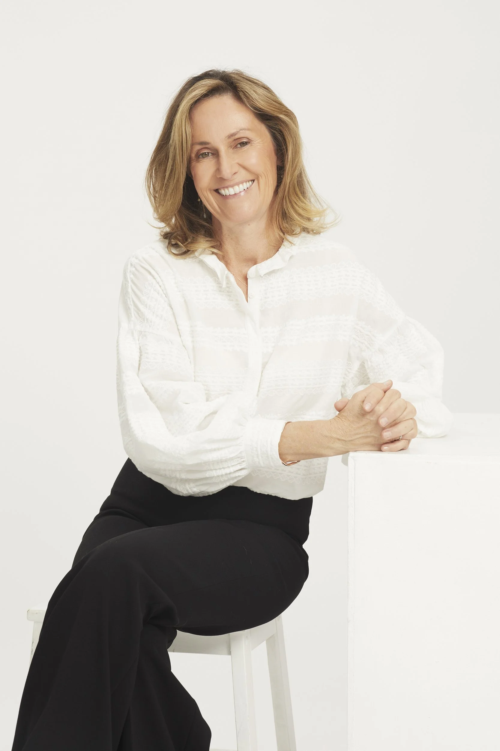 A woman smiling, seated on a stool, wearing a white blouse and black pants against a plain background.