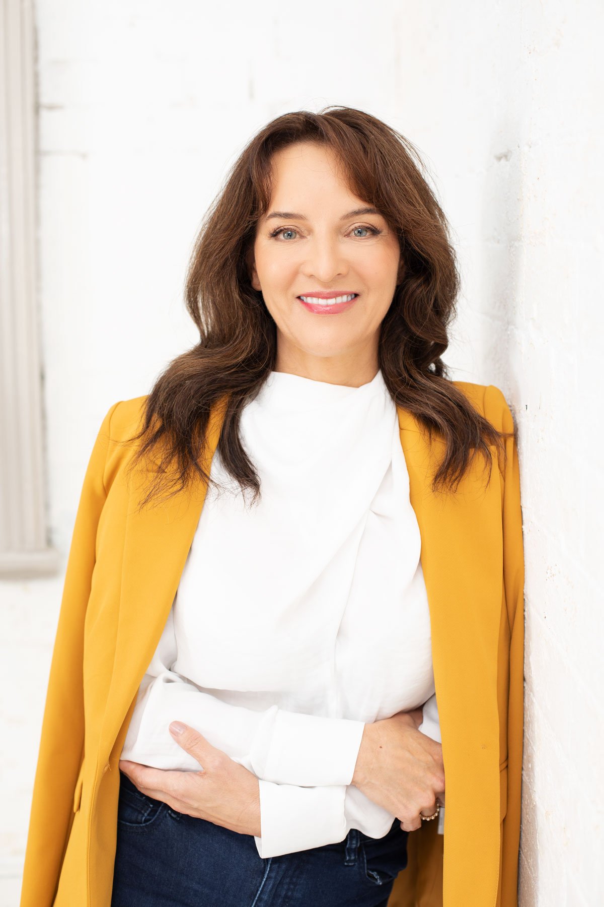 Person wearing a mustard yellow blazer and white blouse, smiling against a white brick wall.