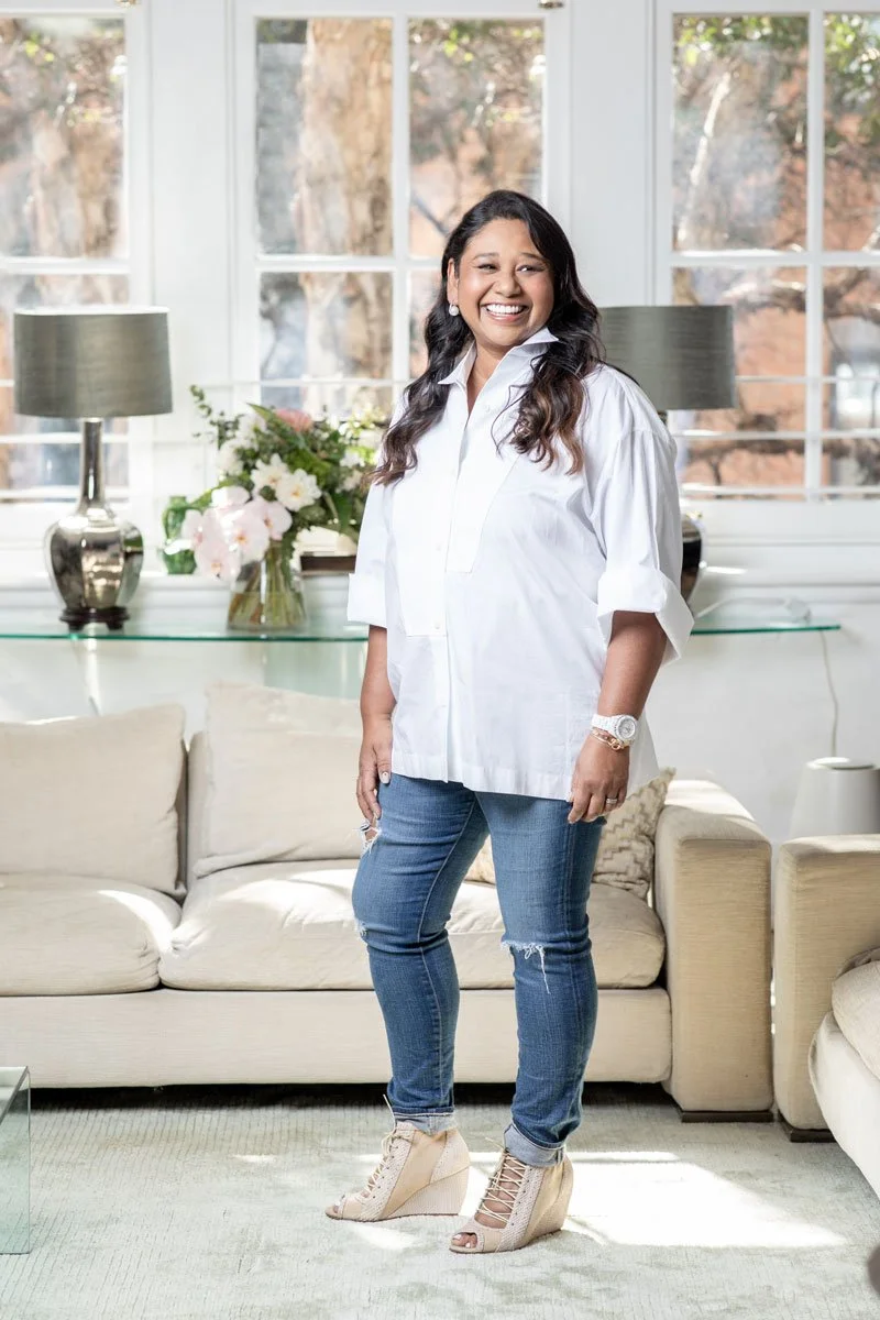 Smiling woman in a white shirt and jeans standing in a living room with a sofa, lamps, and flowers, with sunlight streaming through large windows.