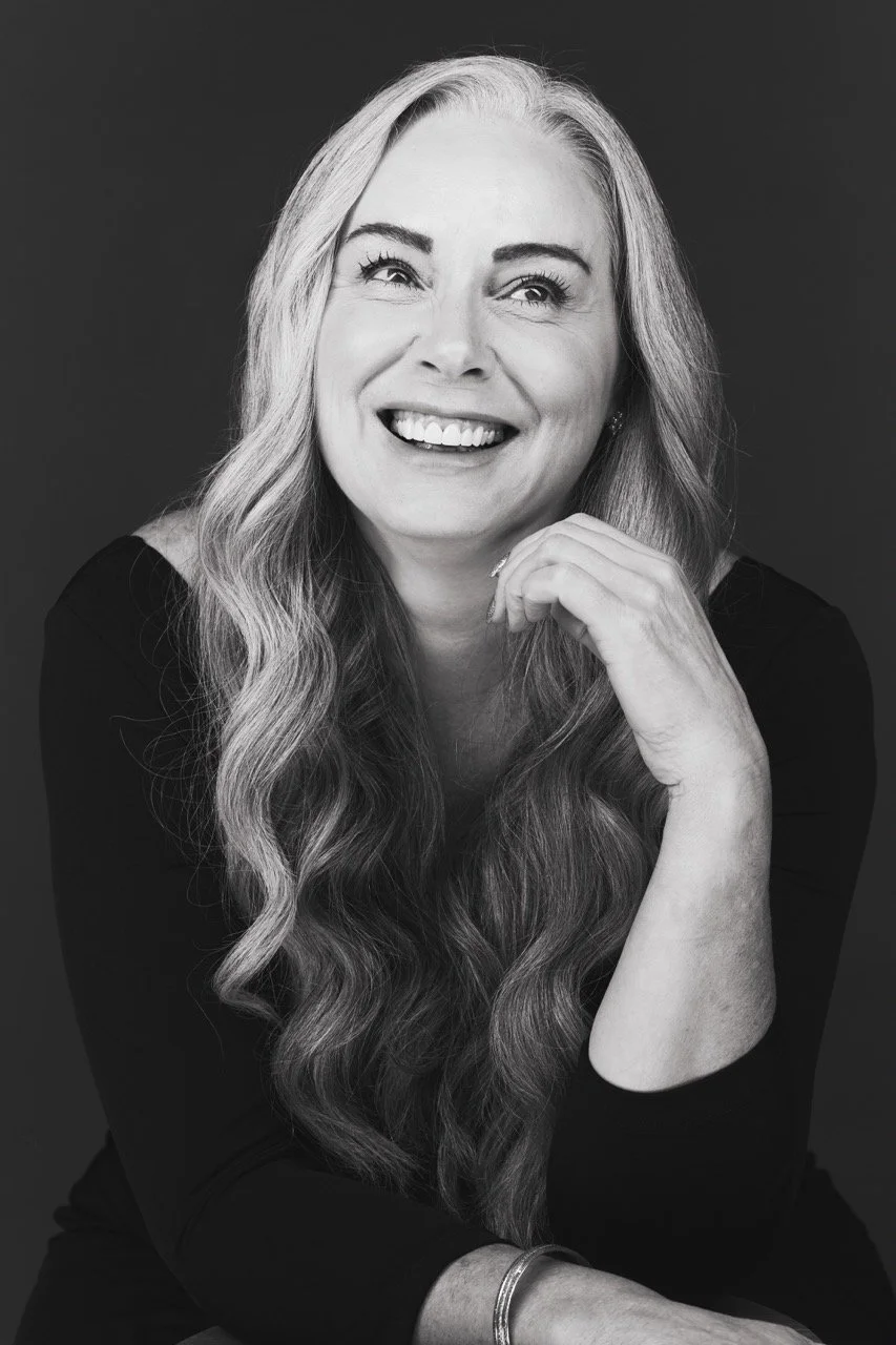 Black and white portrait of a smiling woman with long wavy hair, wearing a dark top and a bracelet.