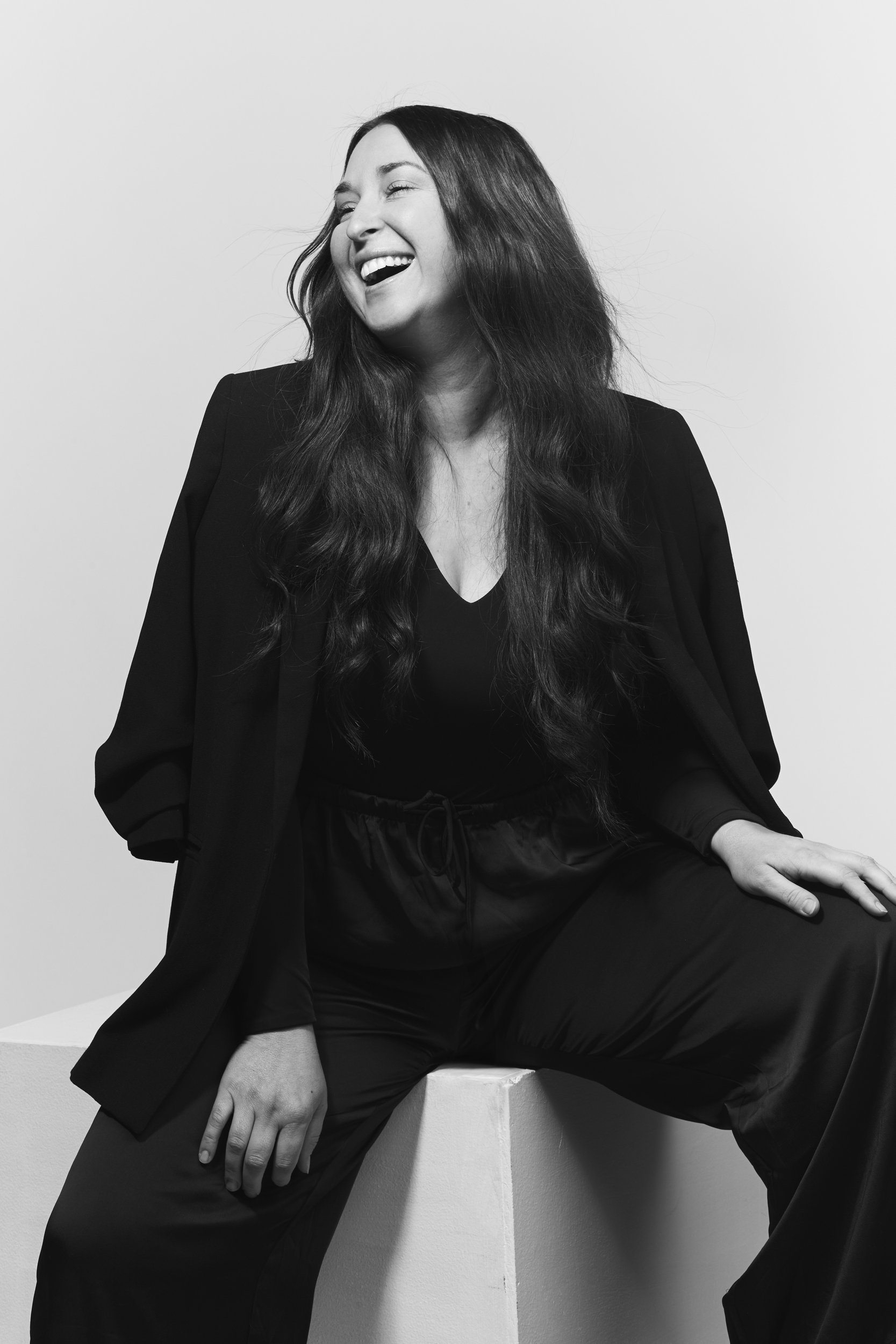 Black and white photo of a woman with long hair and a big smile, sitting on a white cube, wearing a dark outfit against a plain background.