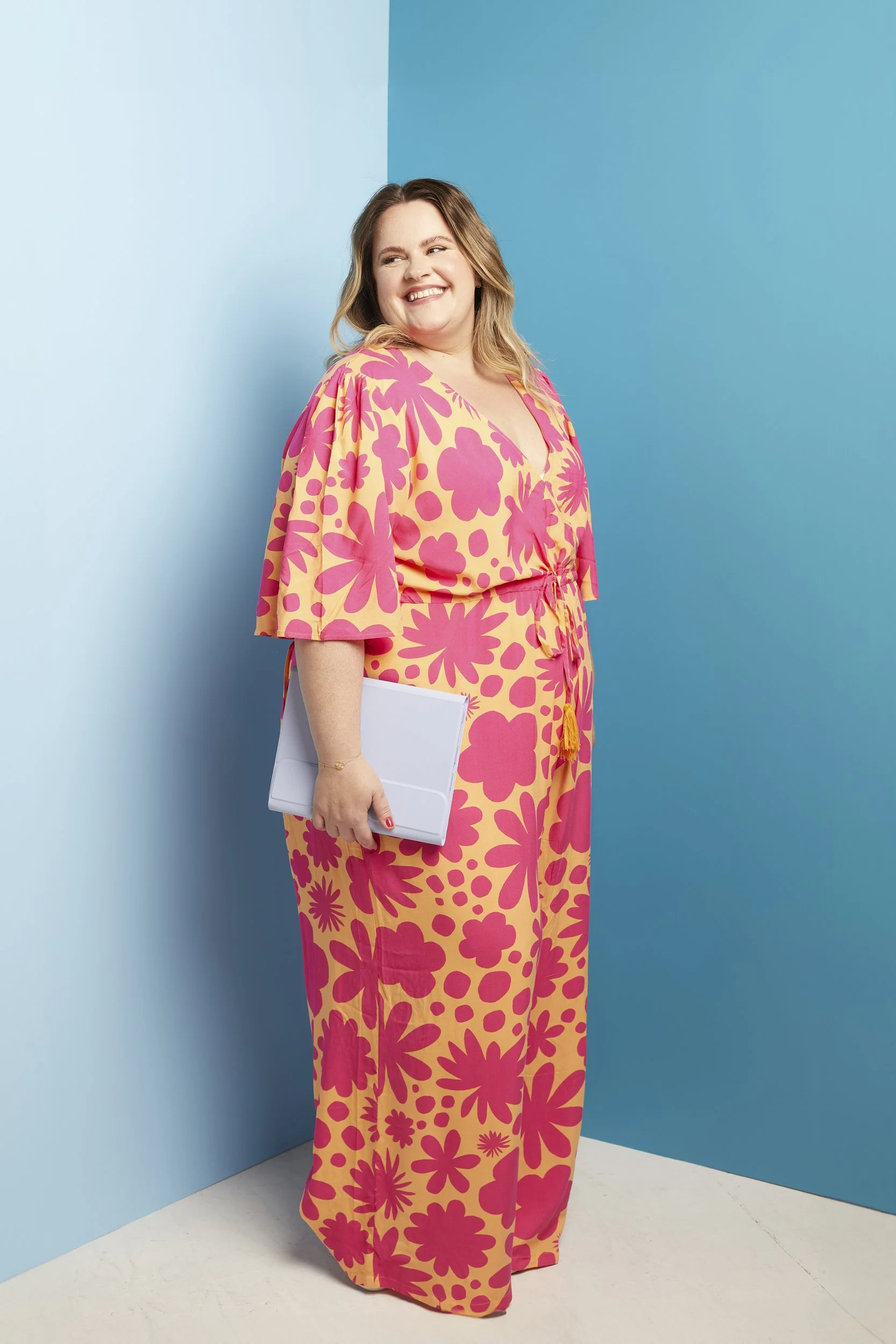 Smiling person in a floral dress holding a white laptop against a blue background.