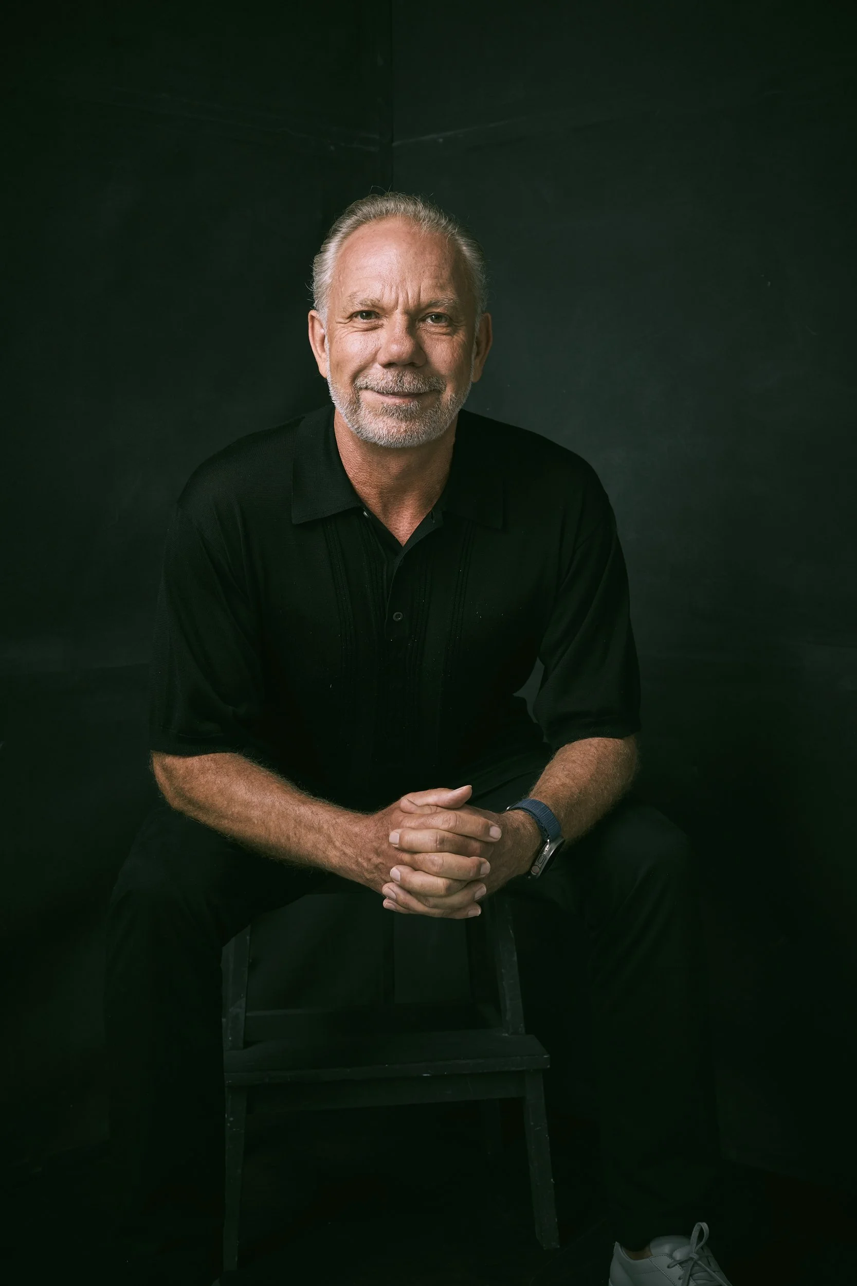 Man sitting on a chair, wearing a black shirt, with hands clasped and smiling slightly.