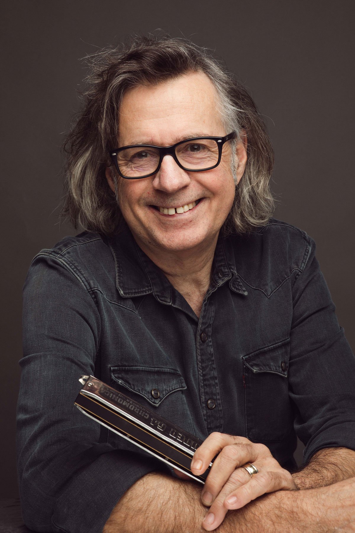 Man with glasses holding a chromatic harmonica, wearing a dark denim shirt, smiling.