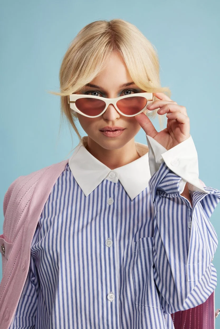 A person with blonde hair wearing sunglasses, a blue and white striped shirt with a white collar, and a pink sweater.