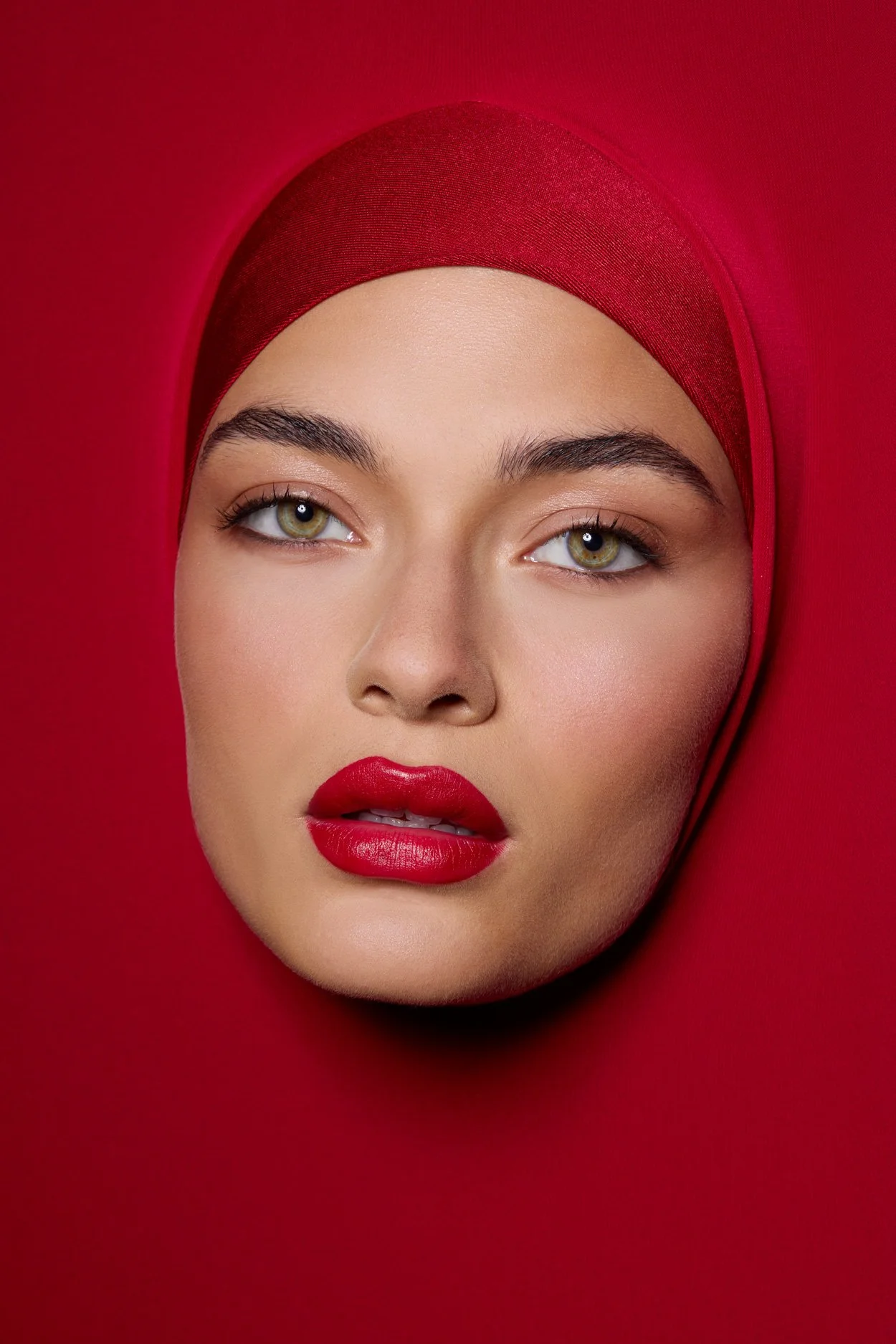 Close-up of a person's face emerging from red fabric, with matching red lipstick and head covering.