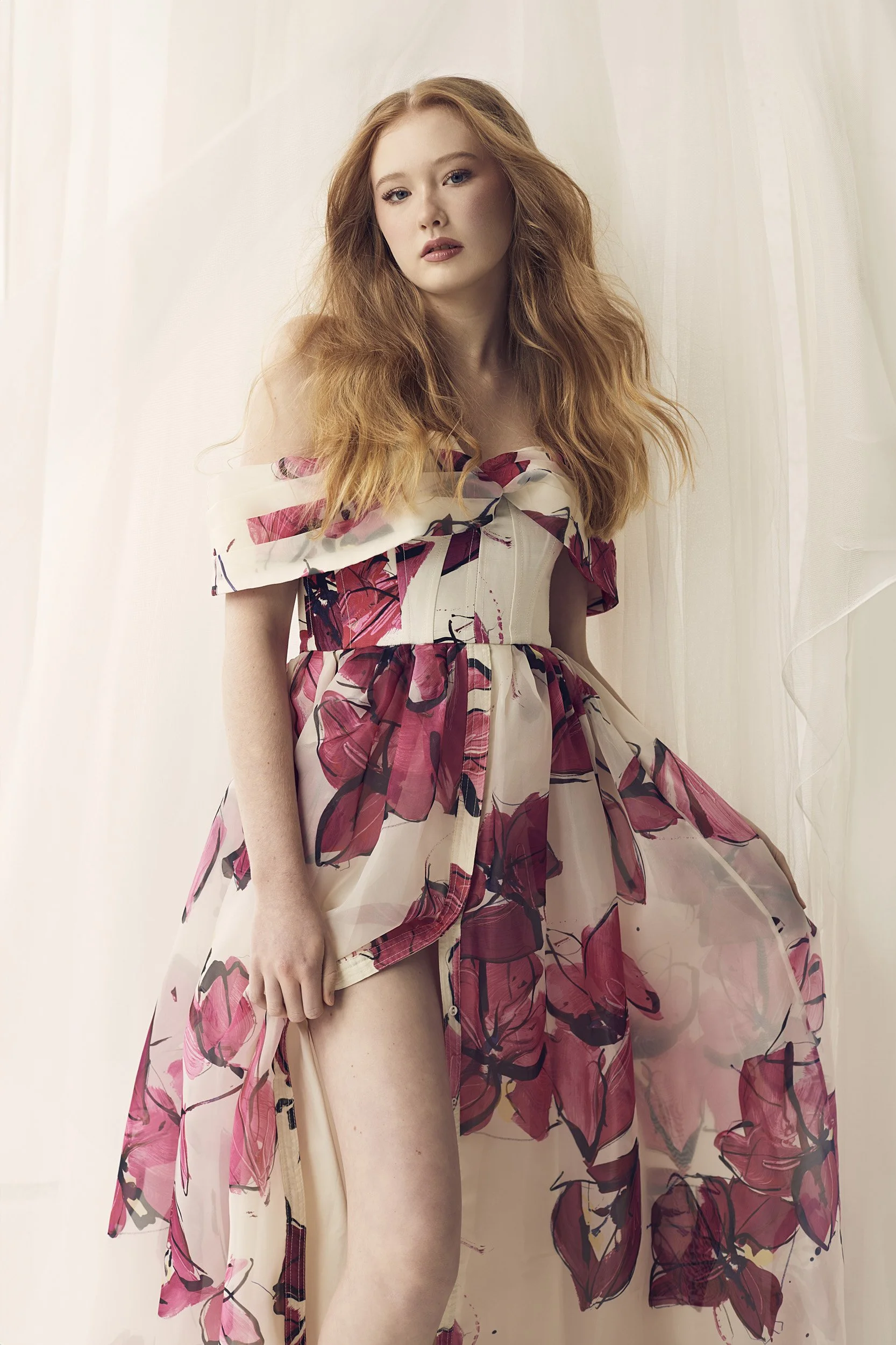 Woman wearing an off-the-shoulder floral dress with pink and red patterns, standing against a light-colored draped background.