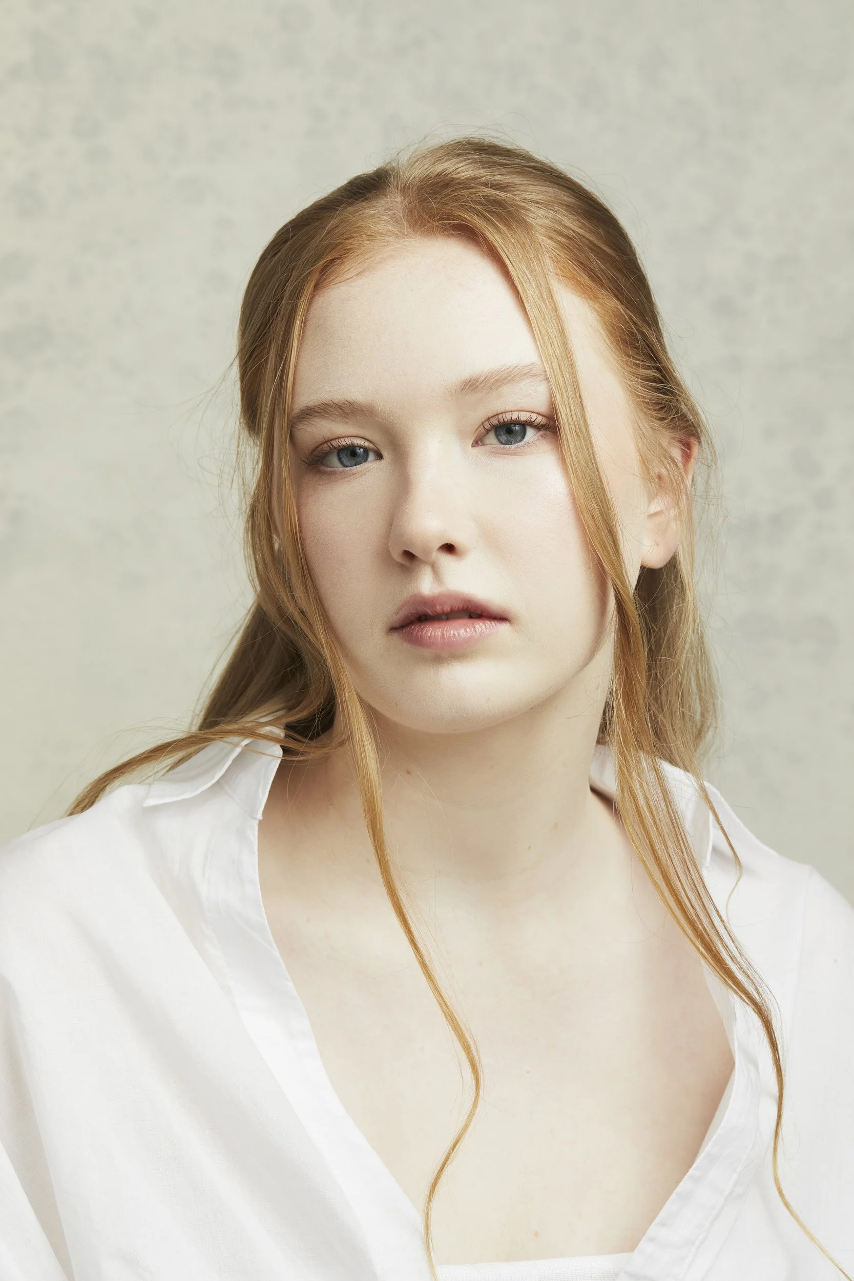 Portrait of a person with long light brown hair wearing a white shirt, looking directly at the camera against a neutral background.