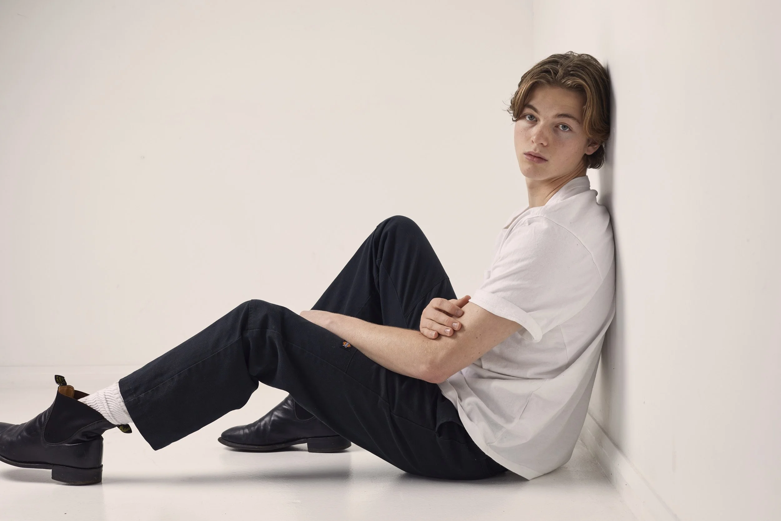 Person sitting on the floor against a white wall, wearing a white t-shirt, black pants, and black ankle boots.