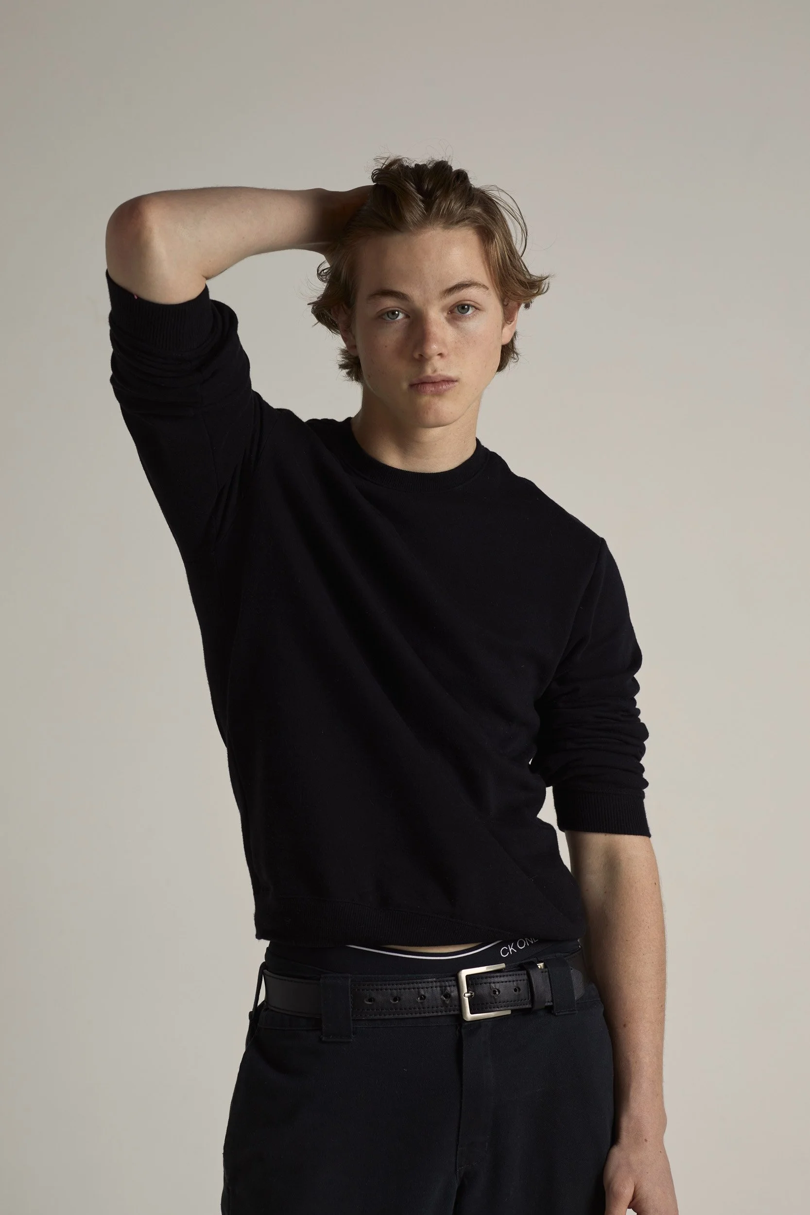 Person wearing black sweater and pants, posing with one hand on head.