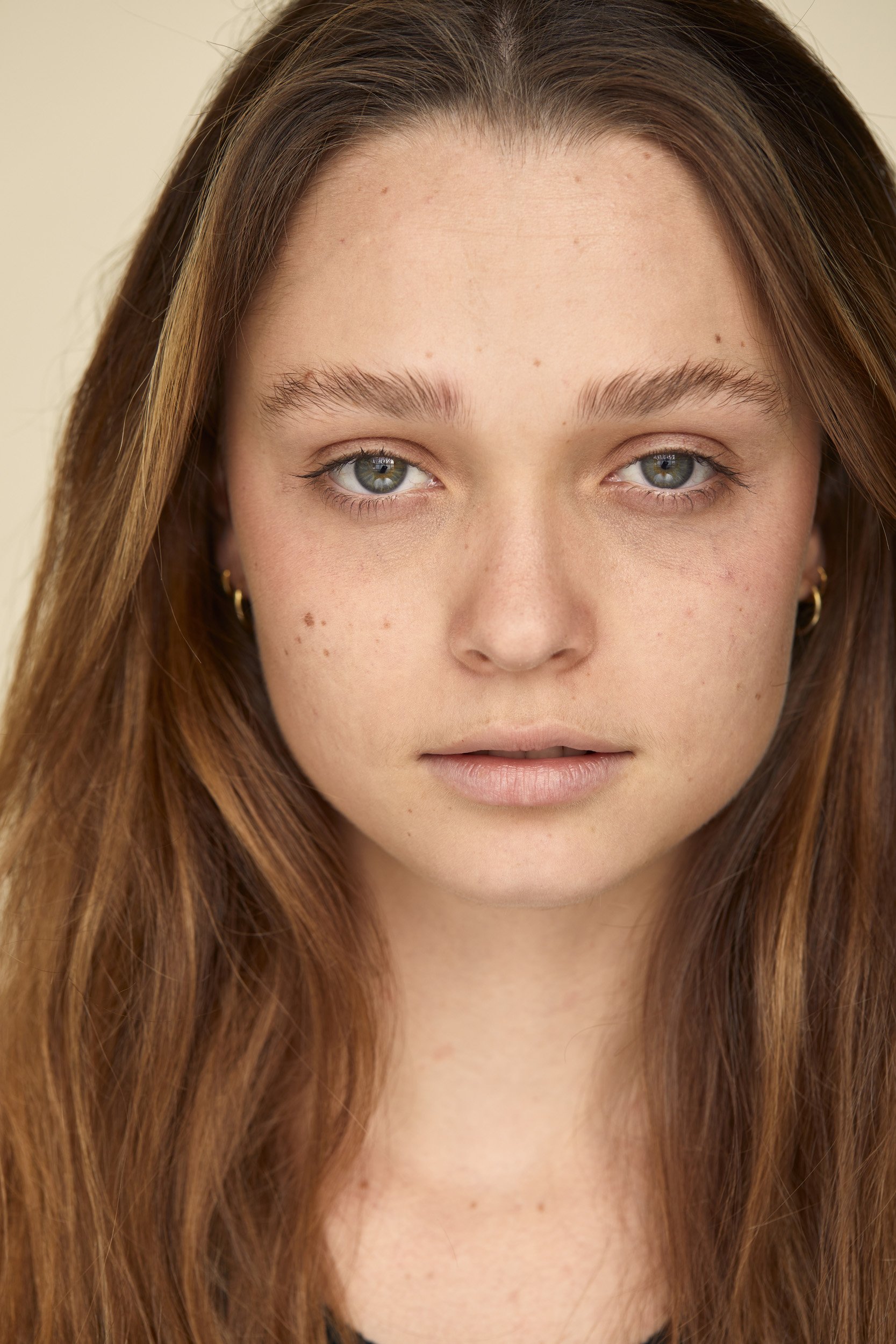 Close-up of a woman's face with long brown hair, natural makeup, and small gold hoop earrings.