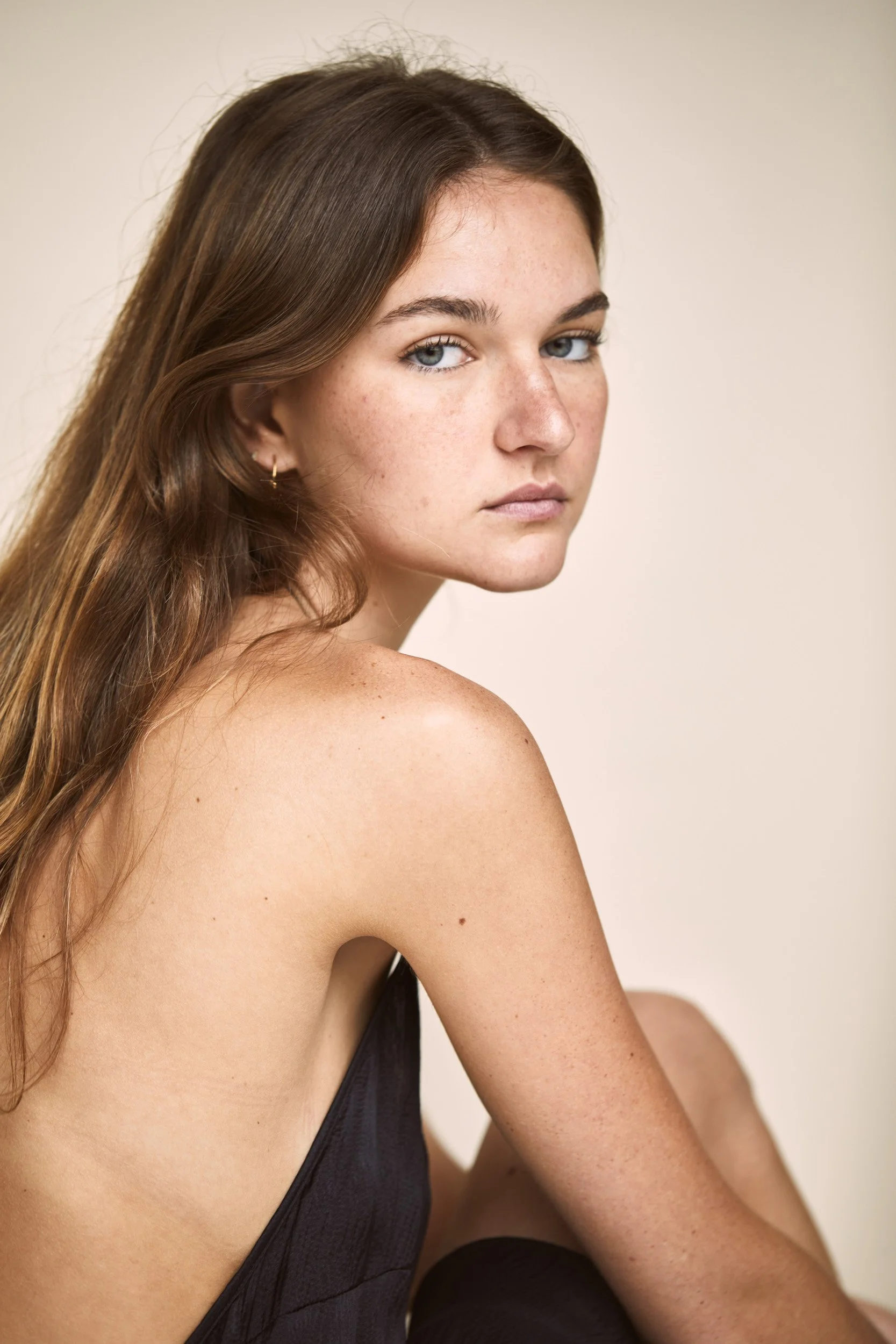 Woman sitting, looking over shoulder, wearing a dark backless dress, neutral background.