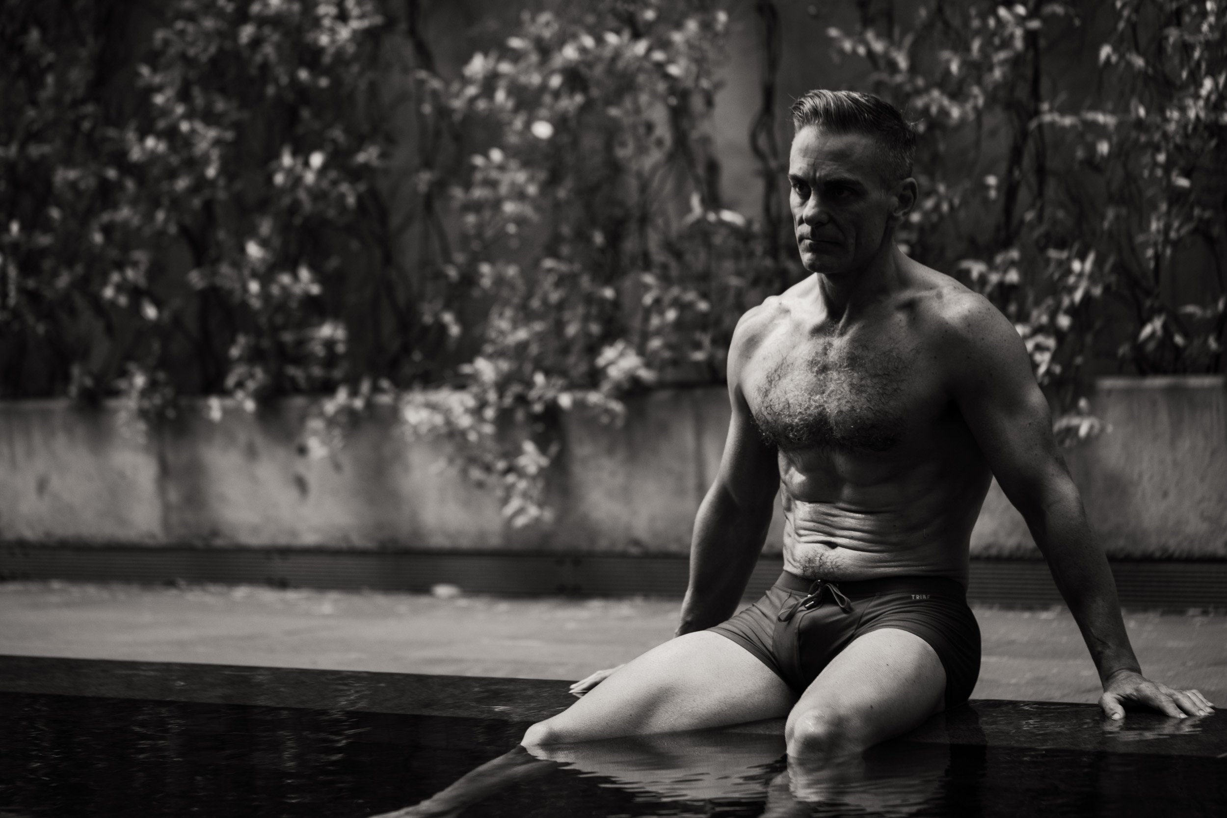 Black and white photo of a muscular man sitting on the edge of a pool with foliage in the background.