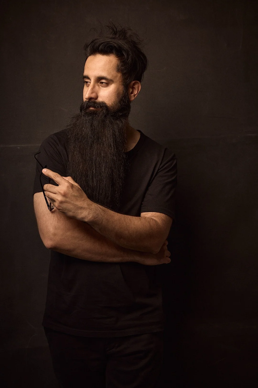 Man with long beard holding eyeglasses, wearing a black t-shirt, against a dark background.