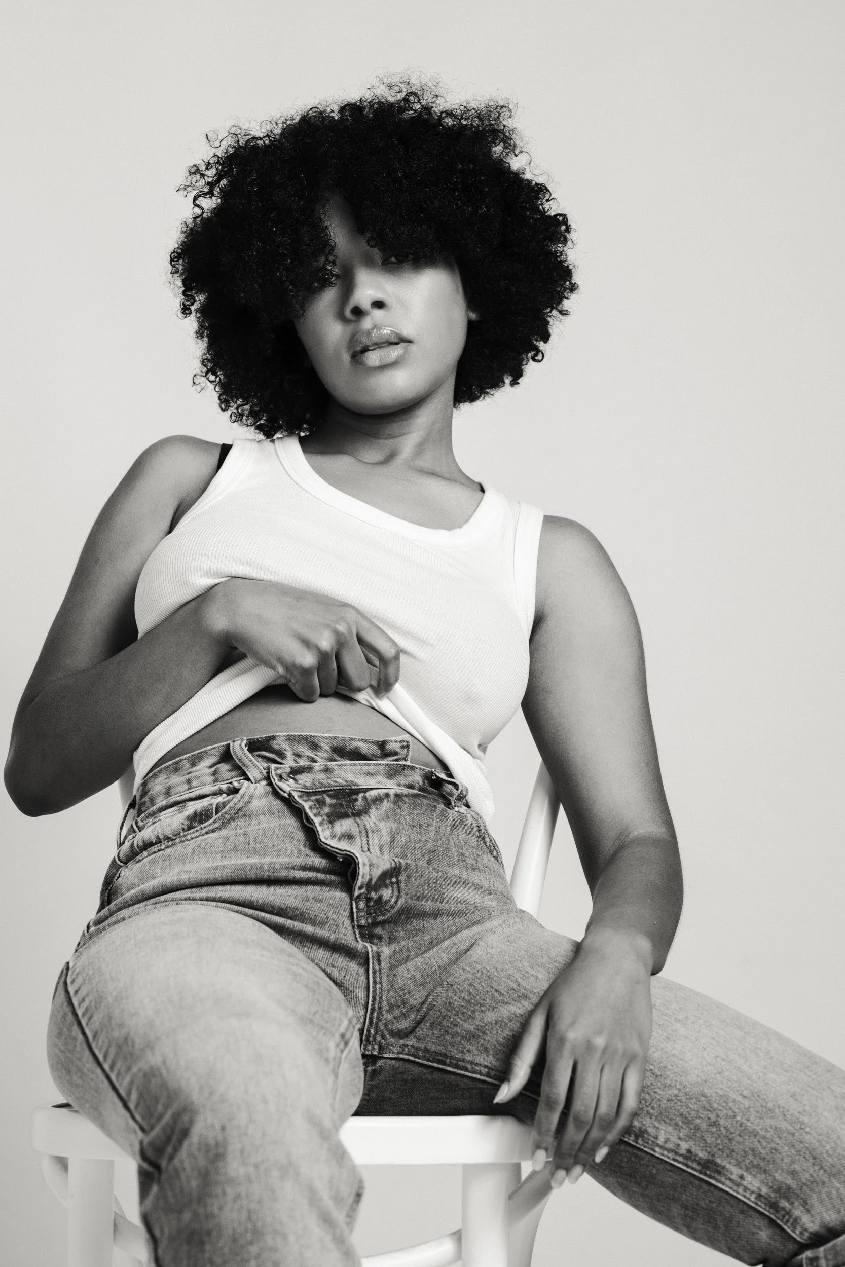 Black and white photo of a woman sitting on a chair, wearing a tank top and jeans, with curly hair and a relaxed expression.