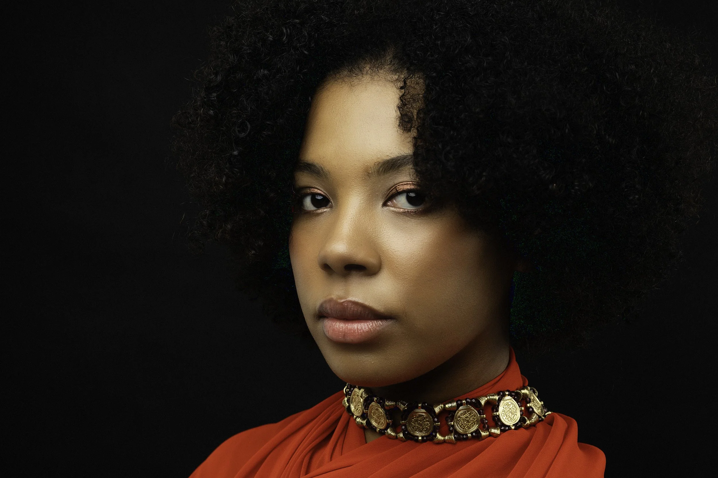Portrait of a woman with curly hair wearing an ornate necklace and an orange top against a dark background.