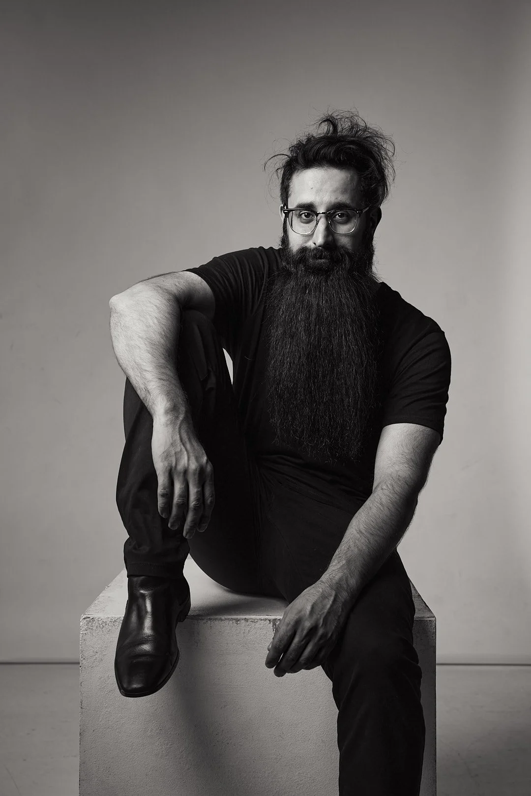Black and white photo of a man with a long beard sitting on a cube, wearing glasses and a black shirt.