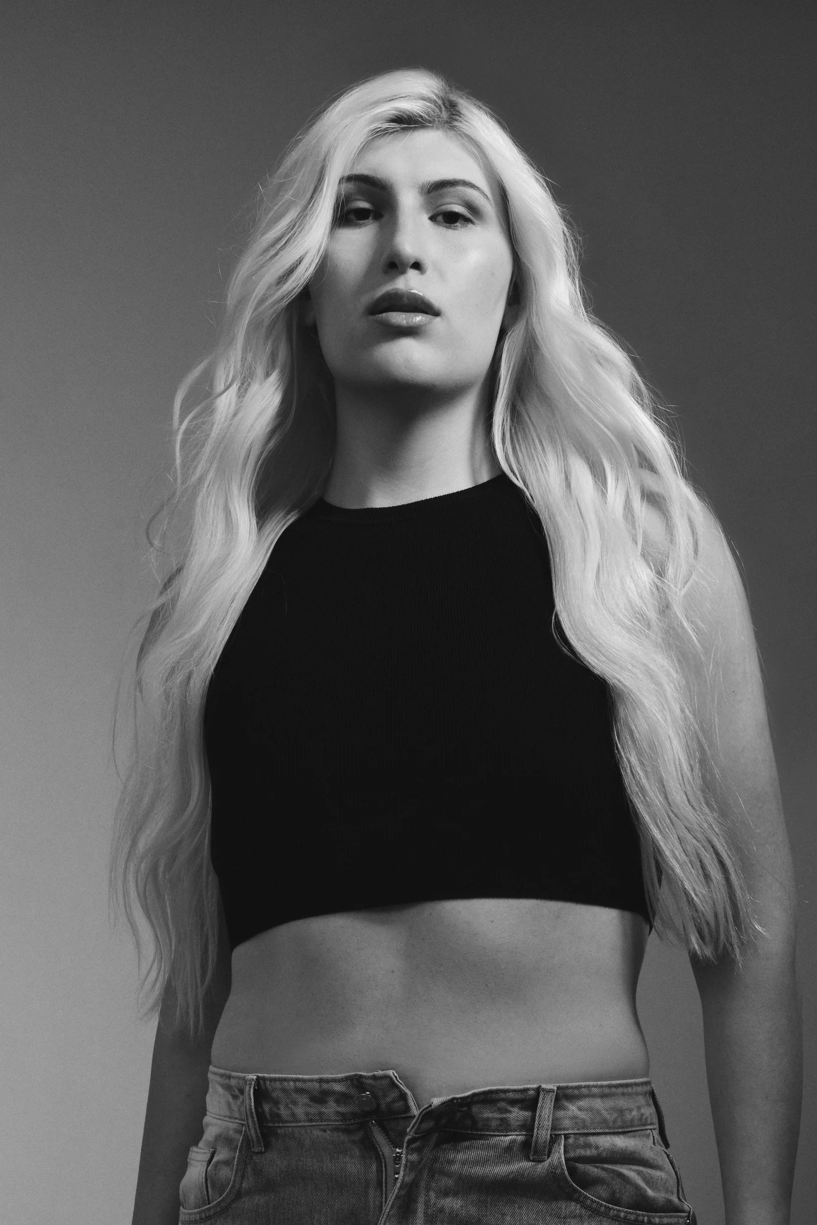 Black and white portrait of a person with long blonde hair wearing a dark crop top and jeans, looking directly at the camera.