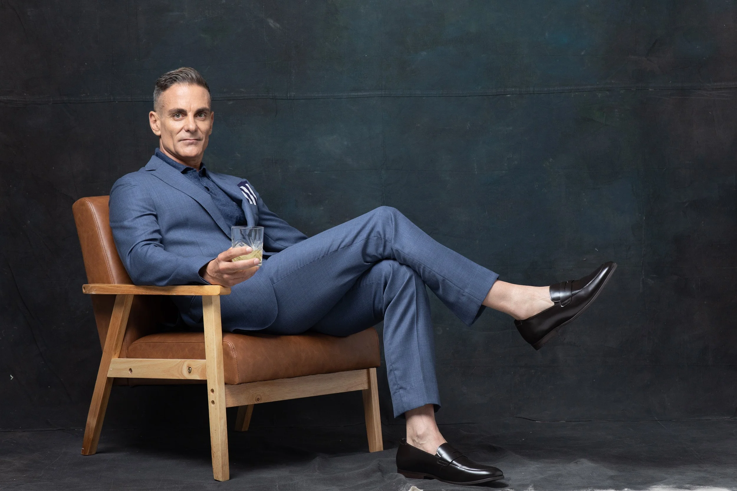 Man in blue suit sitting in chair holding a drink