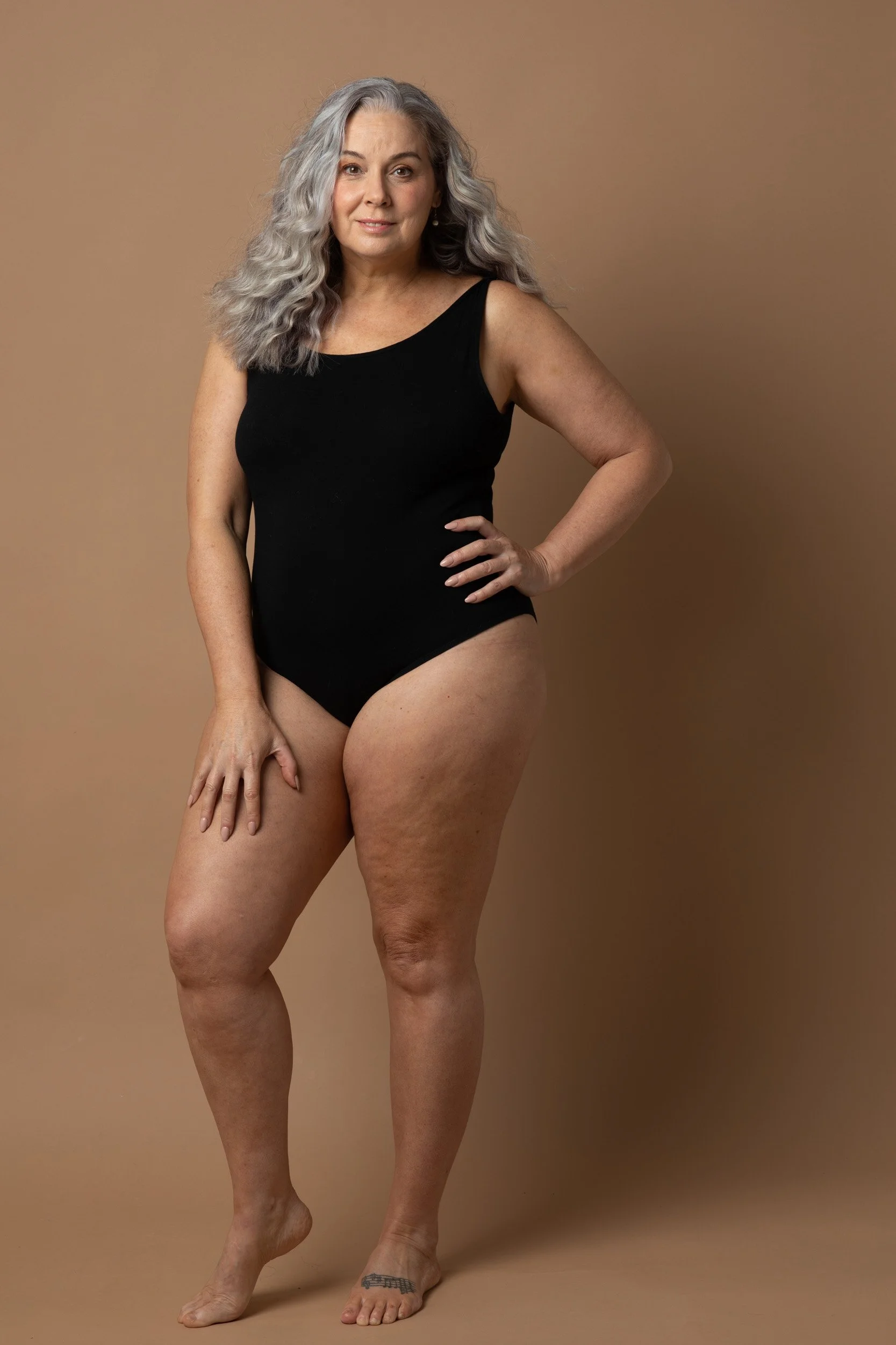 Woman with long gray hair wearing a black swimsuit standing on a brown background.