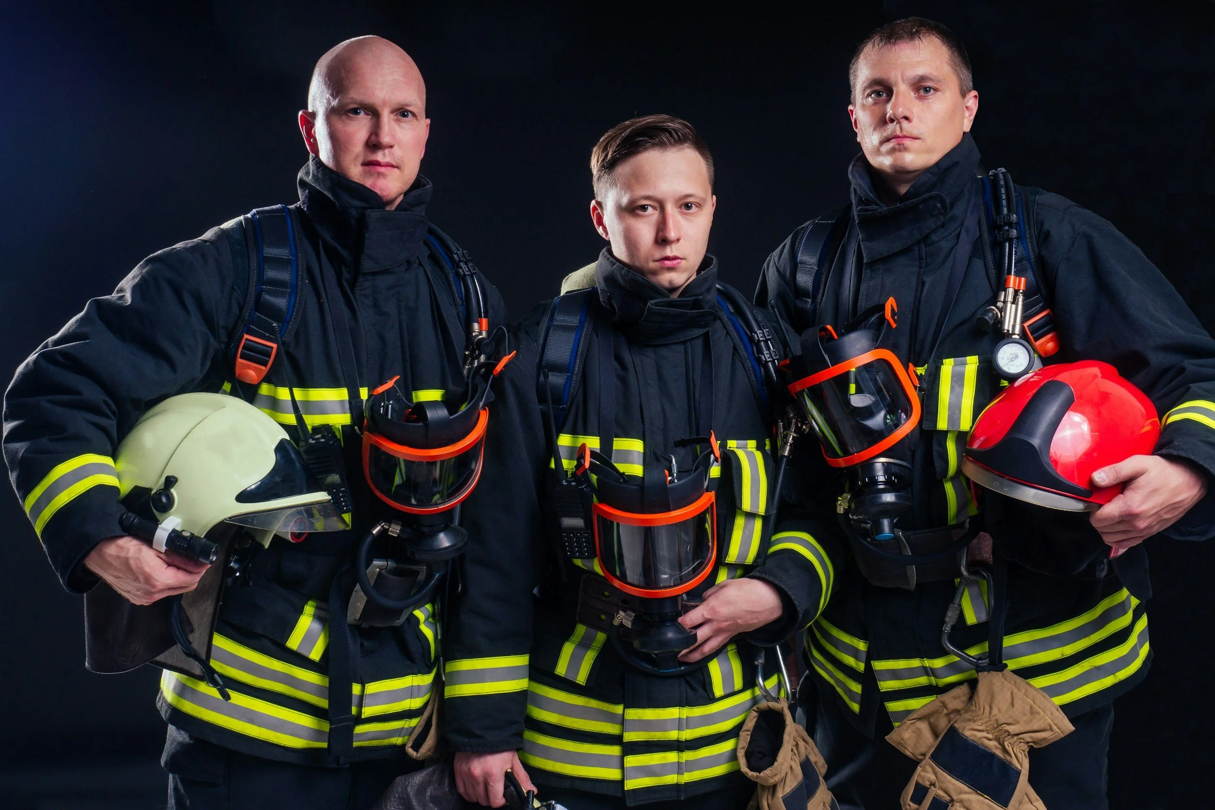 Three male firefighters in black turnout gear with yellow reflective stripes, holding helmets, and carrying various firefighting tools, standing against a dark background.