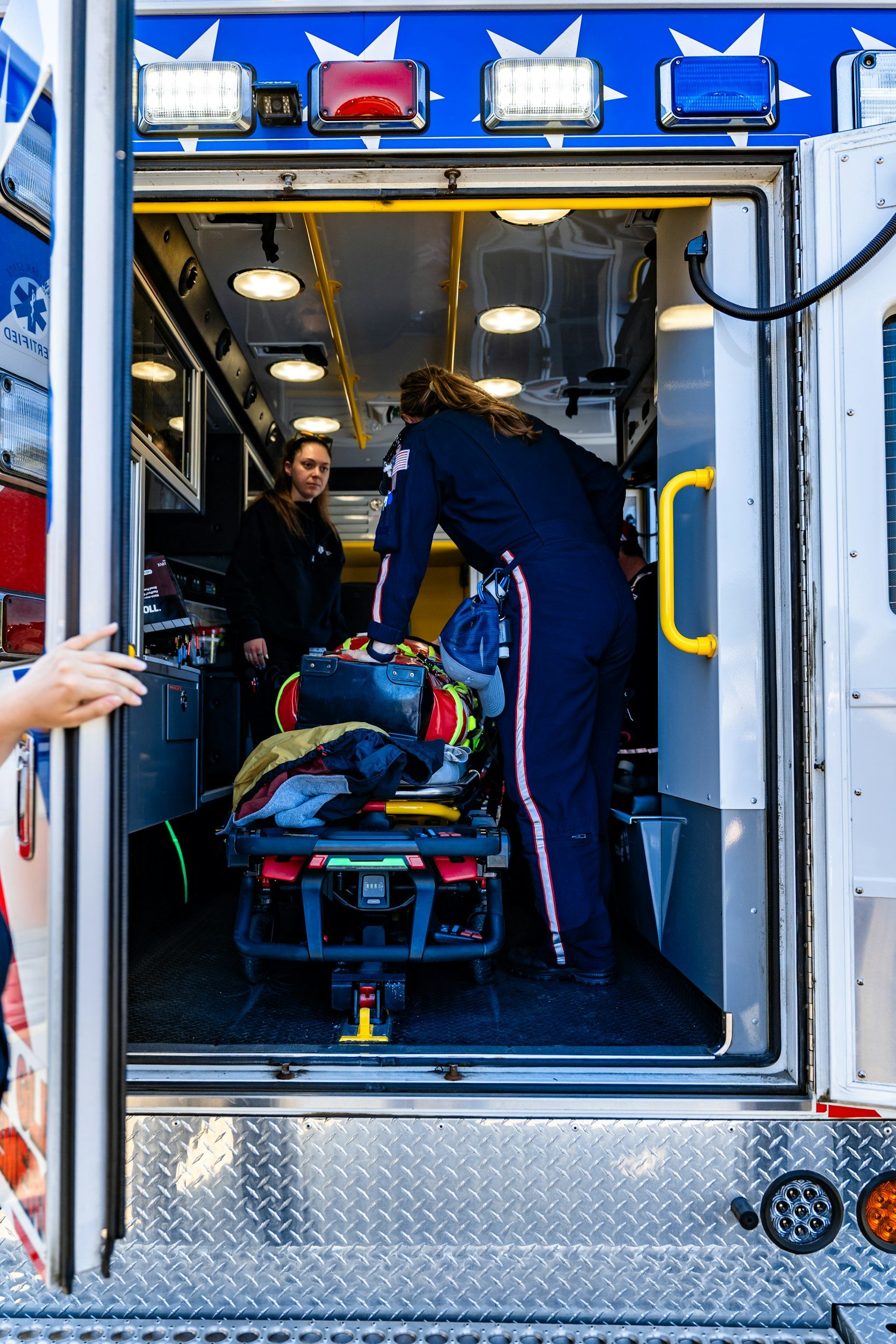 Inside view of an ambulance showing medical personnel preparing equipment, with the ambulance door open, revealing emergency lights and a diamond plate floor.