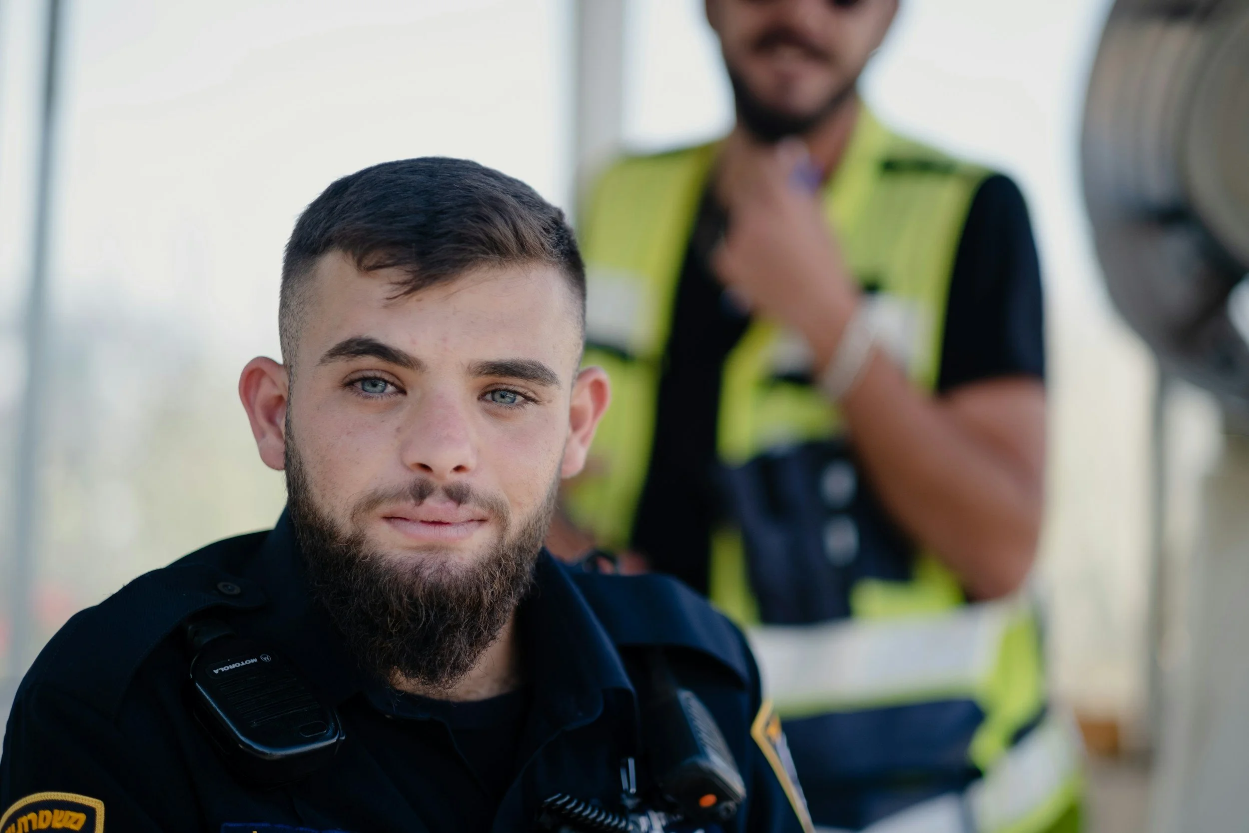 A young man with a beard and blue eyes, wearing a police uniform, is in focus in the foreground. Behind him, a woman in a yellow safety vest and dark shirt, with a blurred face, appears to be inspecting the young man's neck or collar area.