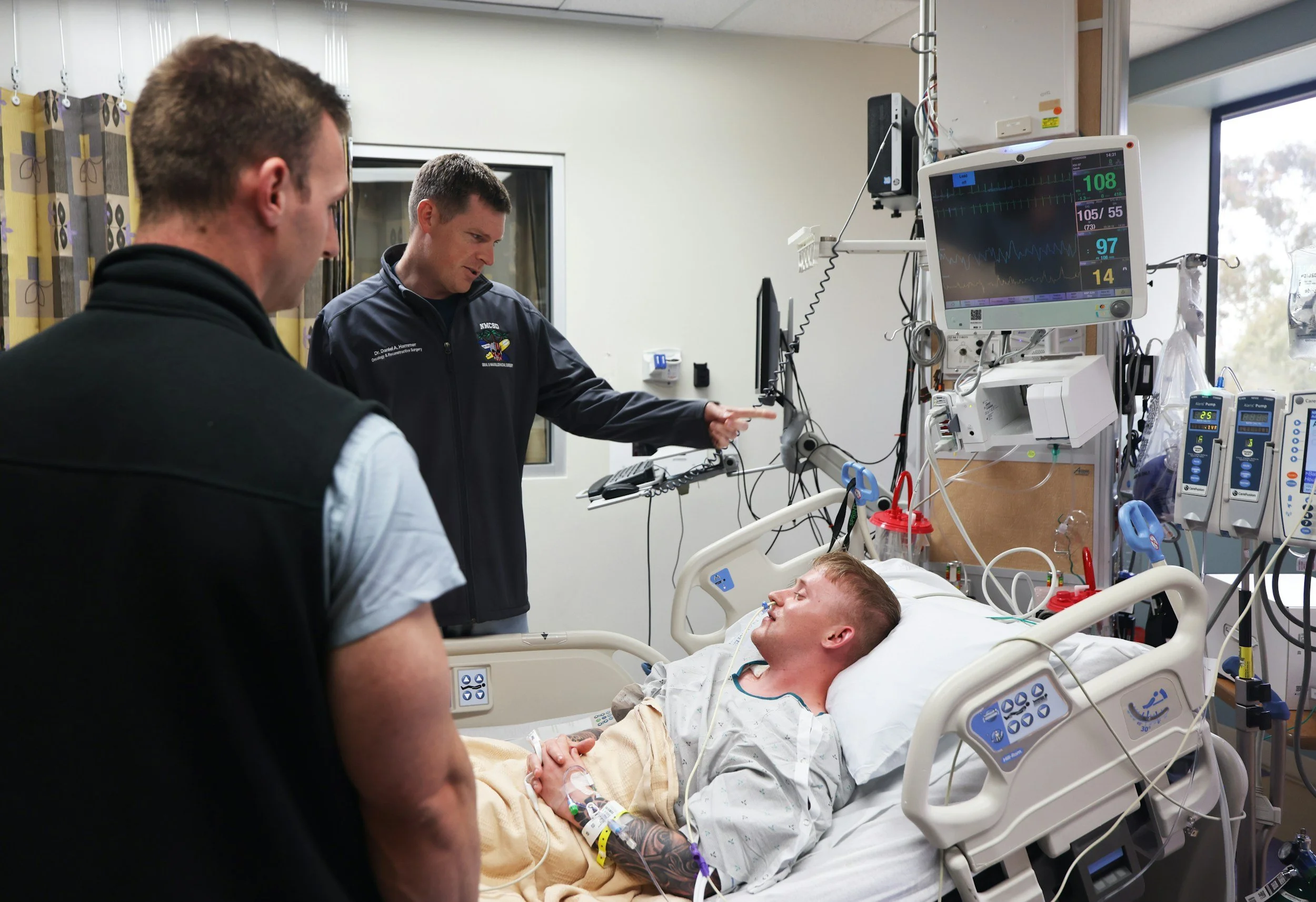 A patient lying in a hospital bed connected to medical monitors and equipment, with two healthcare professionals standing nearby and discussing the patient.