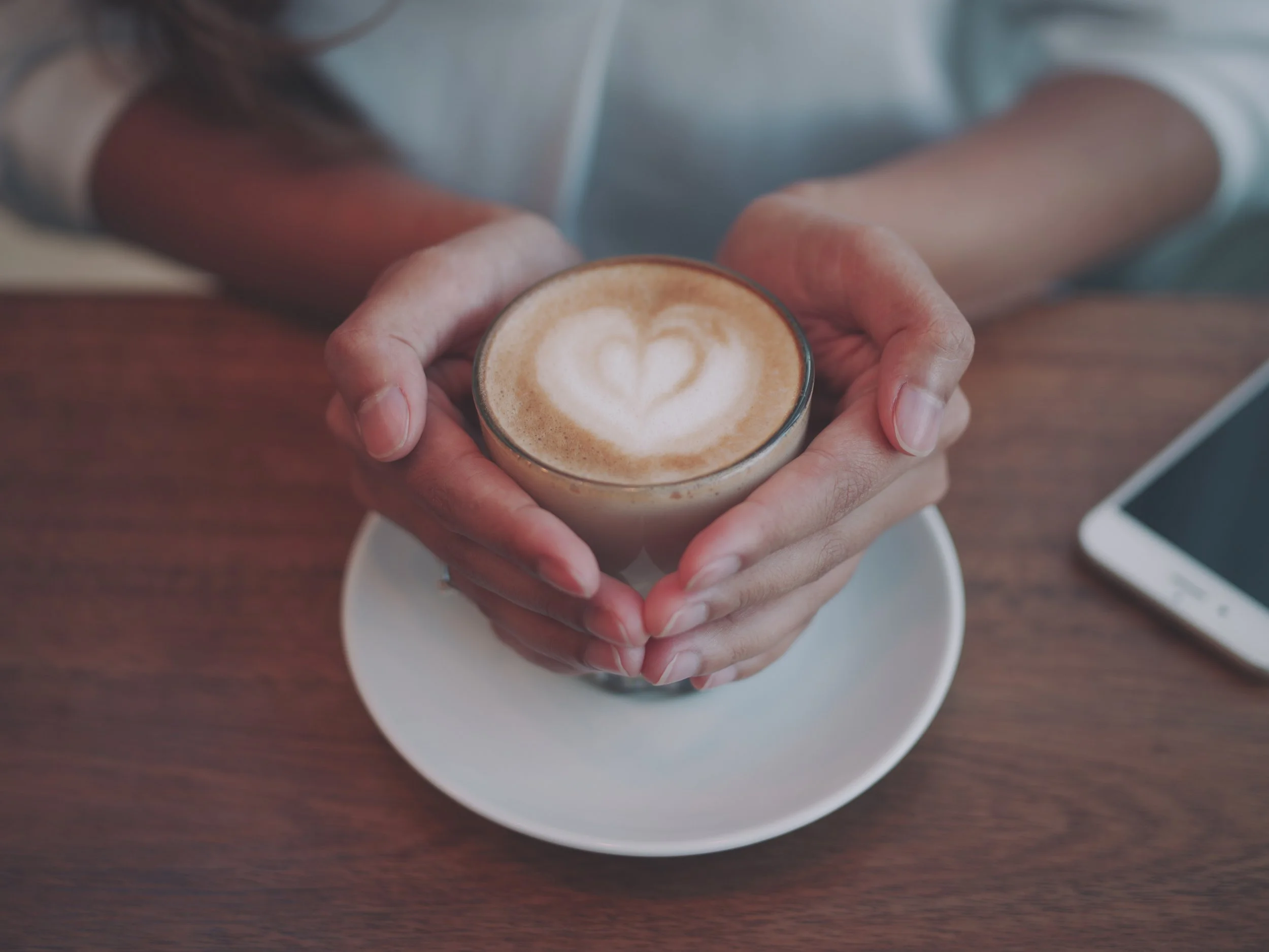 A person holding a small glass of cappuccino with heart-shaped latte art, sitting at a wooden table.
