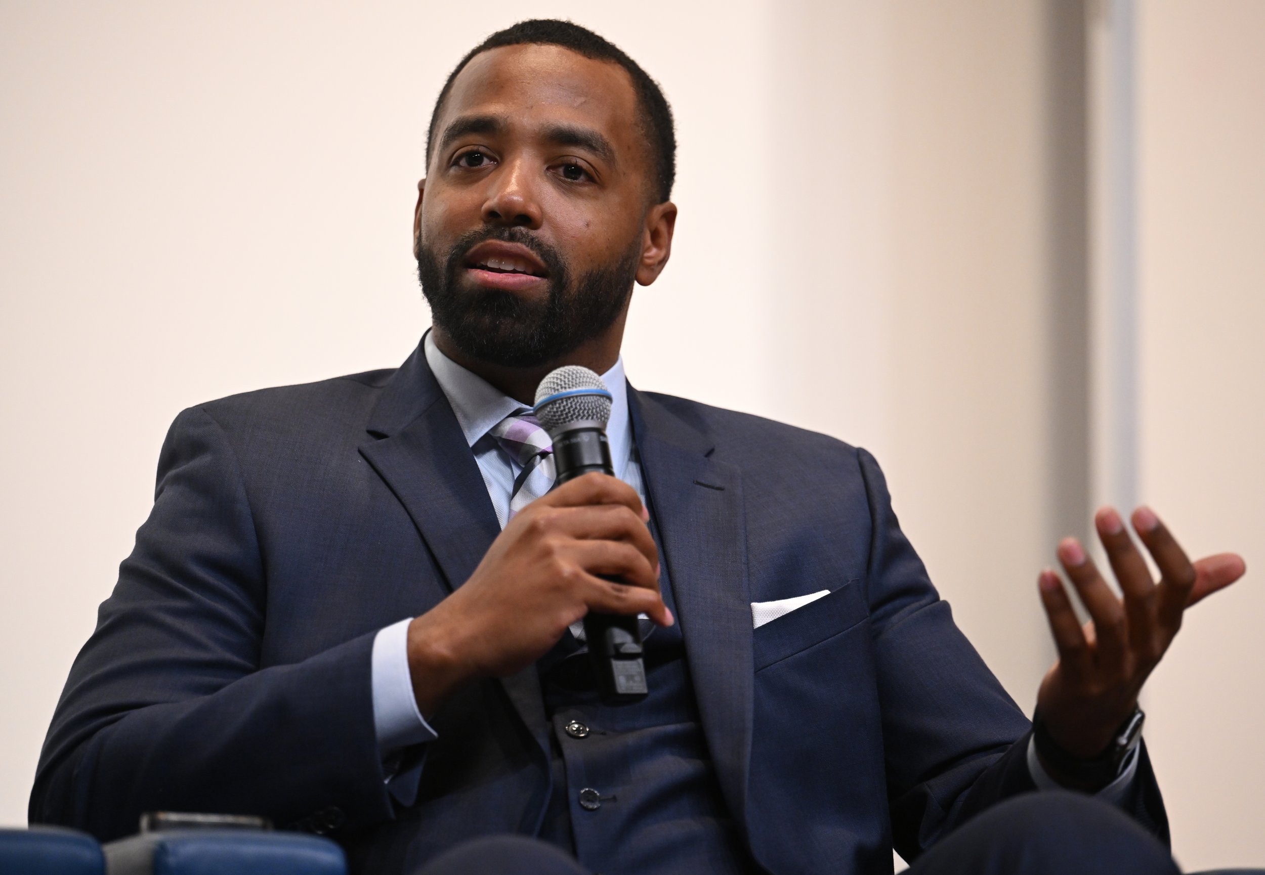 A man in a suit speaking into a microphone while seated.