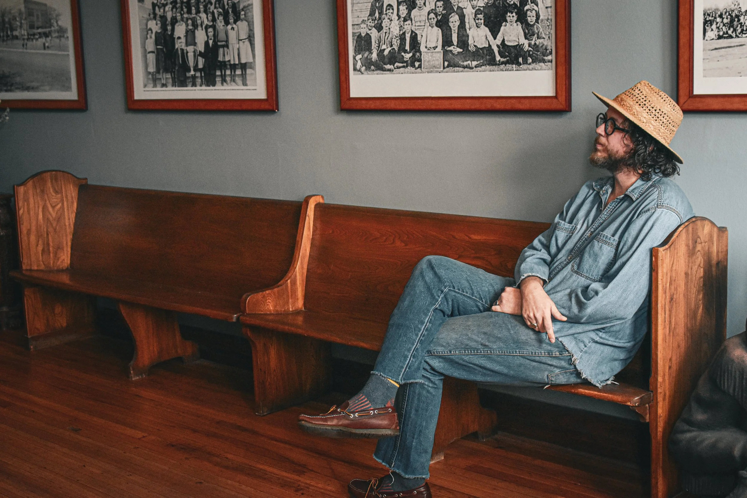 A man with long curly hair, beard, and big glasses sitting on a wooden bench in a room with vintage black and white photographs on the wall. He is wearing a straw hat, denim shirt, jeans, and brown boots with red socks.