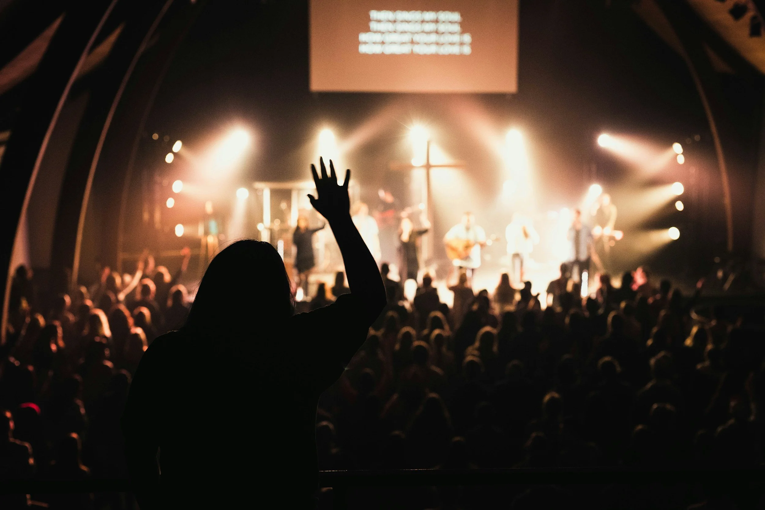 A person in the audience raising their hand during a concert or church service with a band or choir performing on stage in the background, illuminated by stage lights.