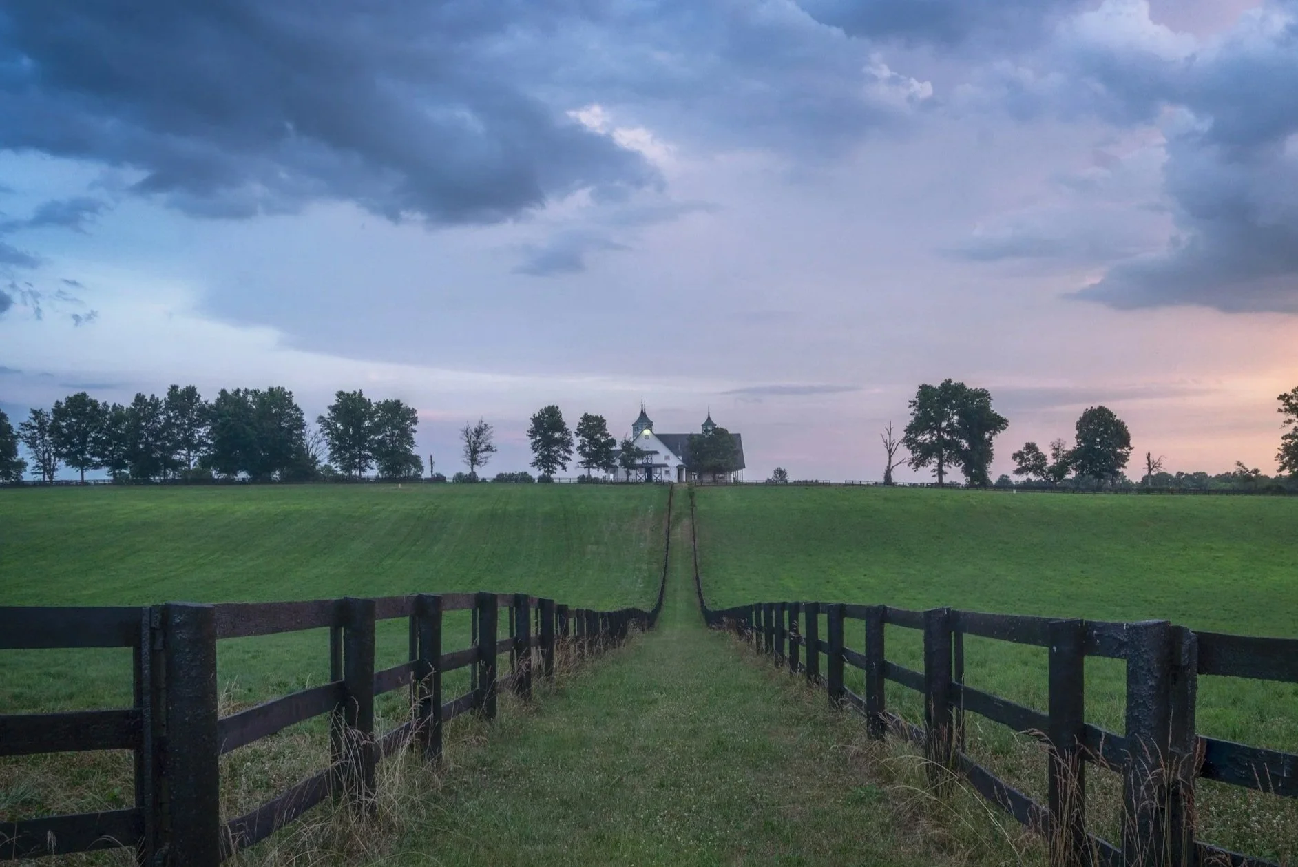 A rural landscape with a fence leading to a church on a hill, surrounded by trees under cloudy skies during sunset or sunrise.